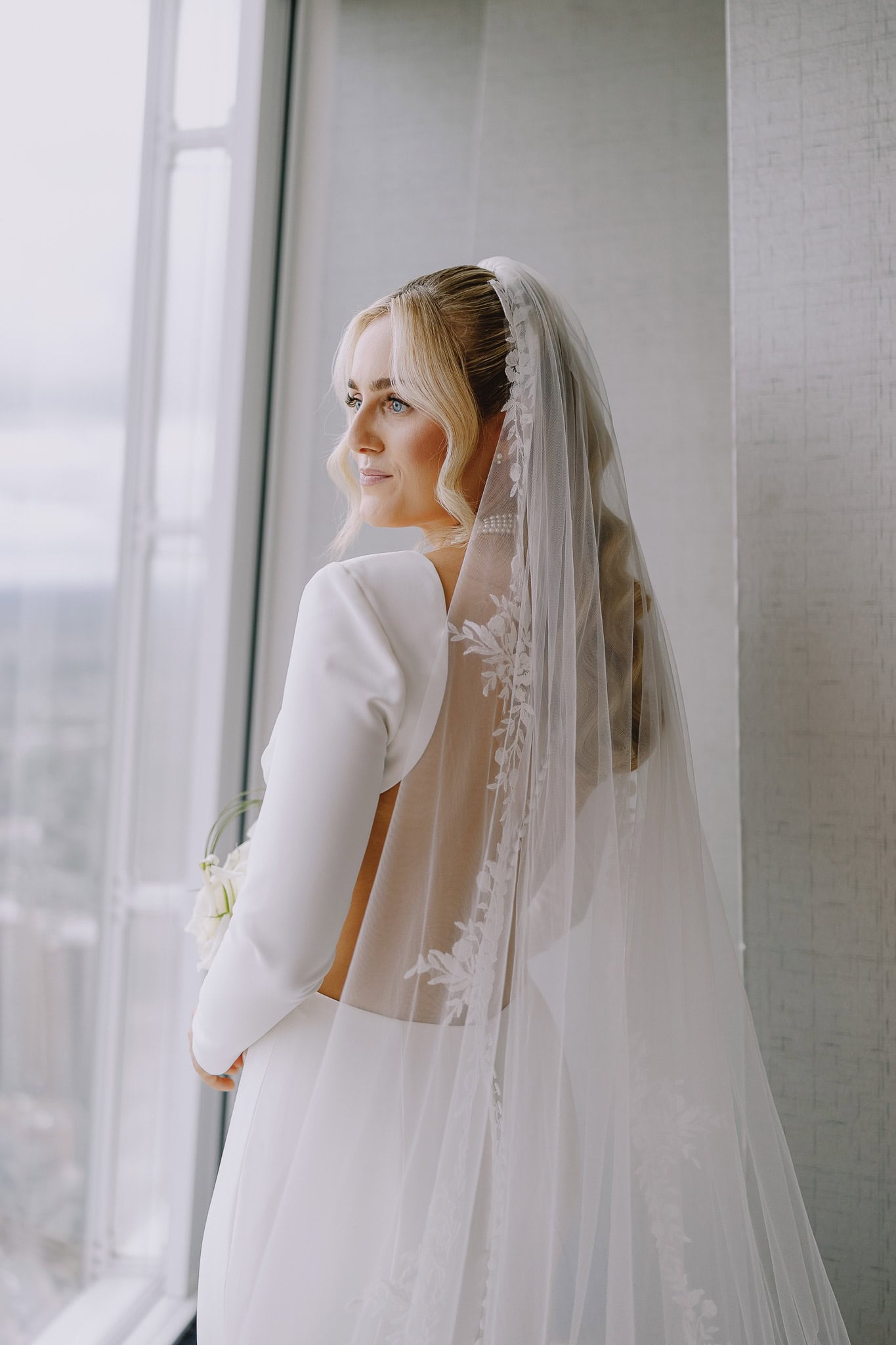 Bride in elegant white dress and veil looks out window, part of wedding photography portfolio.