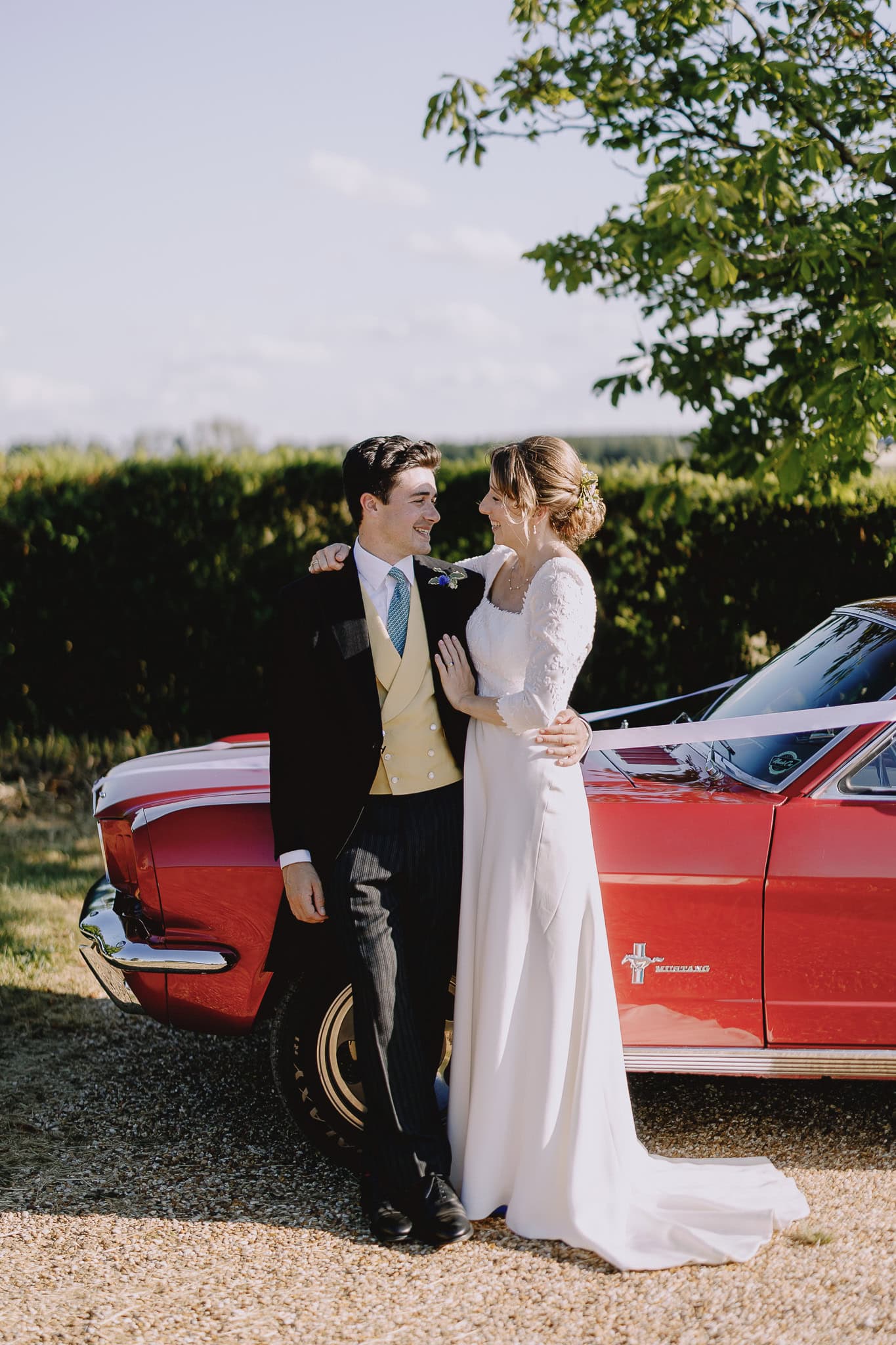 Wedding photo: Bride and groom pose by a red Mustang. Wedding photography portfolio.