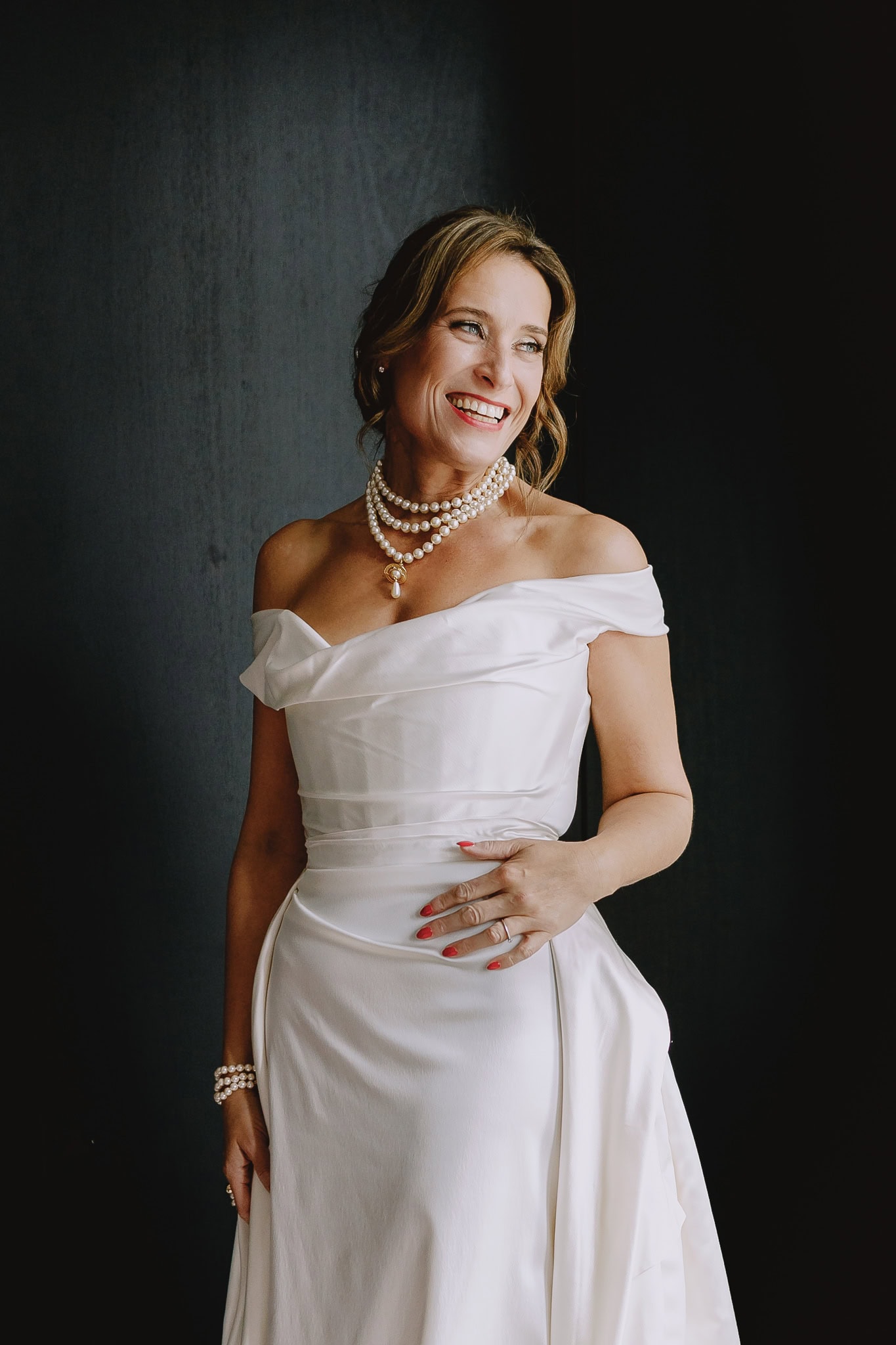 Woman in white off-the-shoulder dress and pearl necklace smiling. Editorial wedding photography.