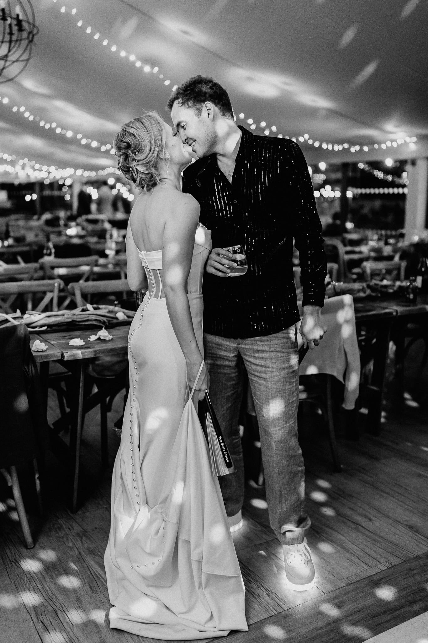 Bride and groom share a kiss, editorial wedding photography style, under string lights.