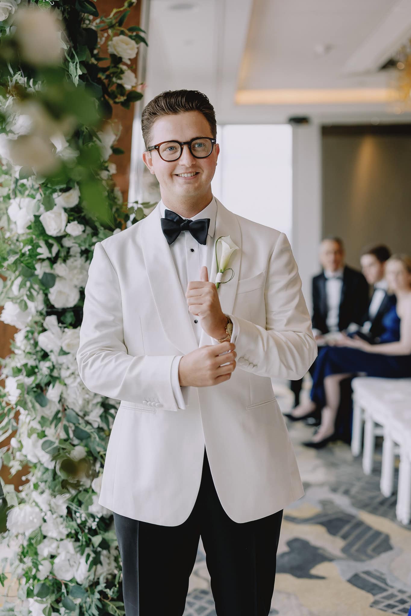 Groom in a white tuxedo and black bow tie at an editorial wedding photography event, smiling confidently.