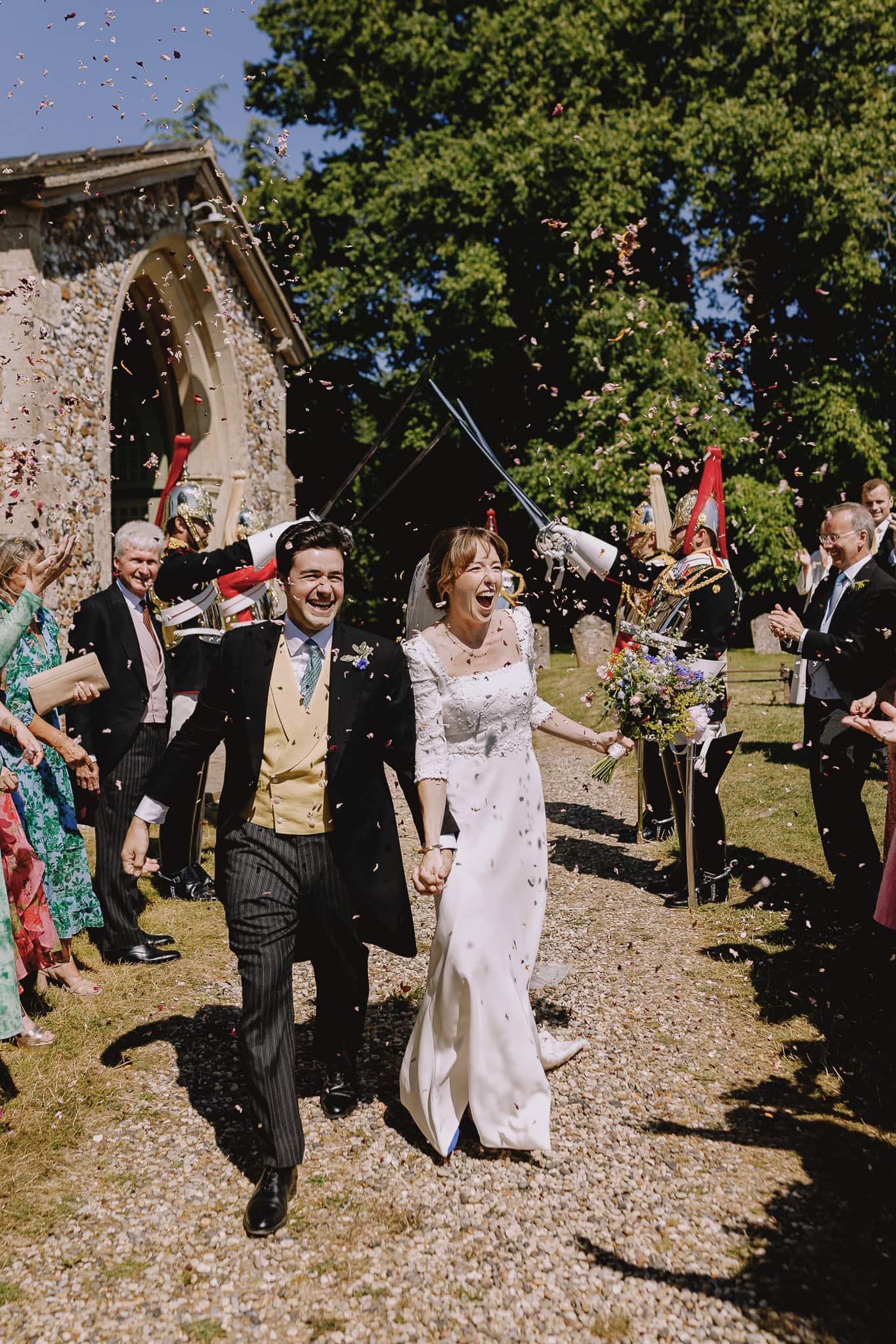 Joyful bride and groom exiting church under sword arch, showered with confetti. Editorial wedding photography.