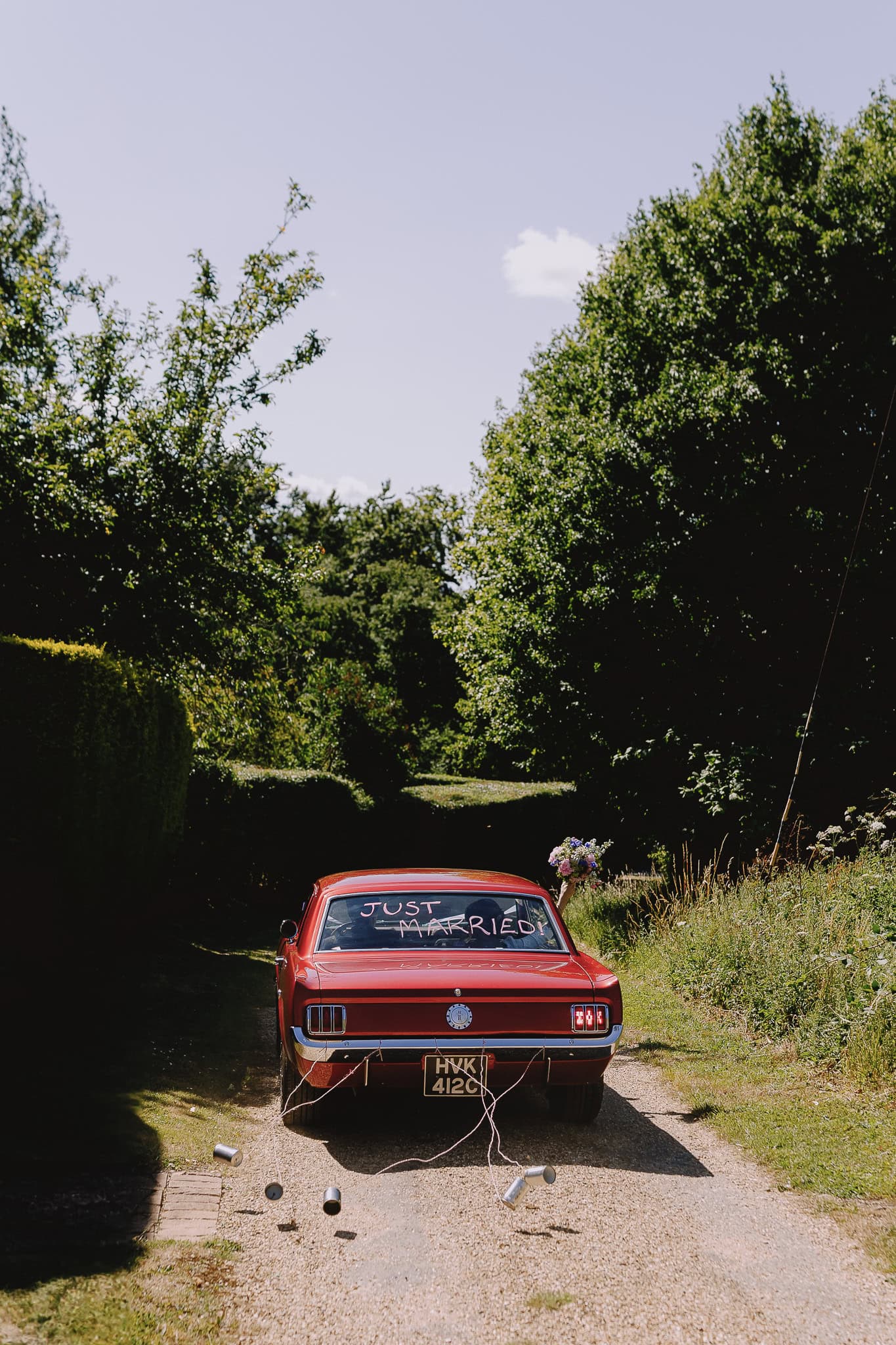 Red vintage car with "Just Married" written on the back, cans tied to the bumper. Editorial wedding photography.