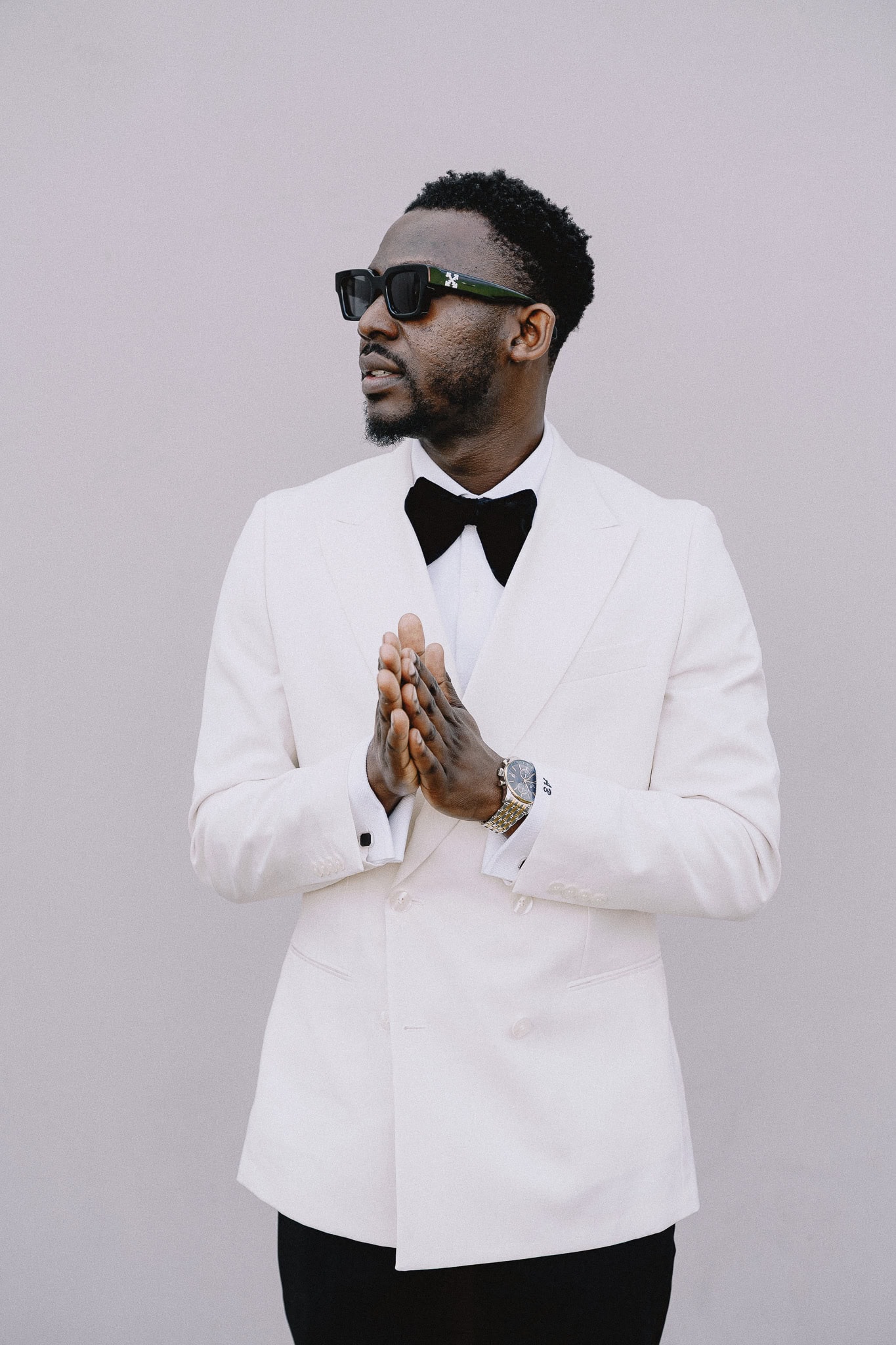Man in white suit with bow tie and sunglasses, hands clasped.