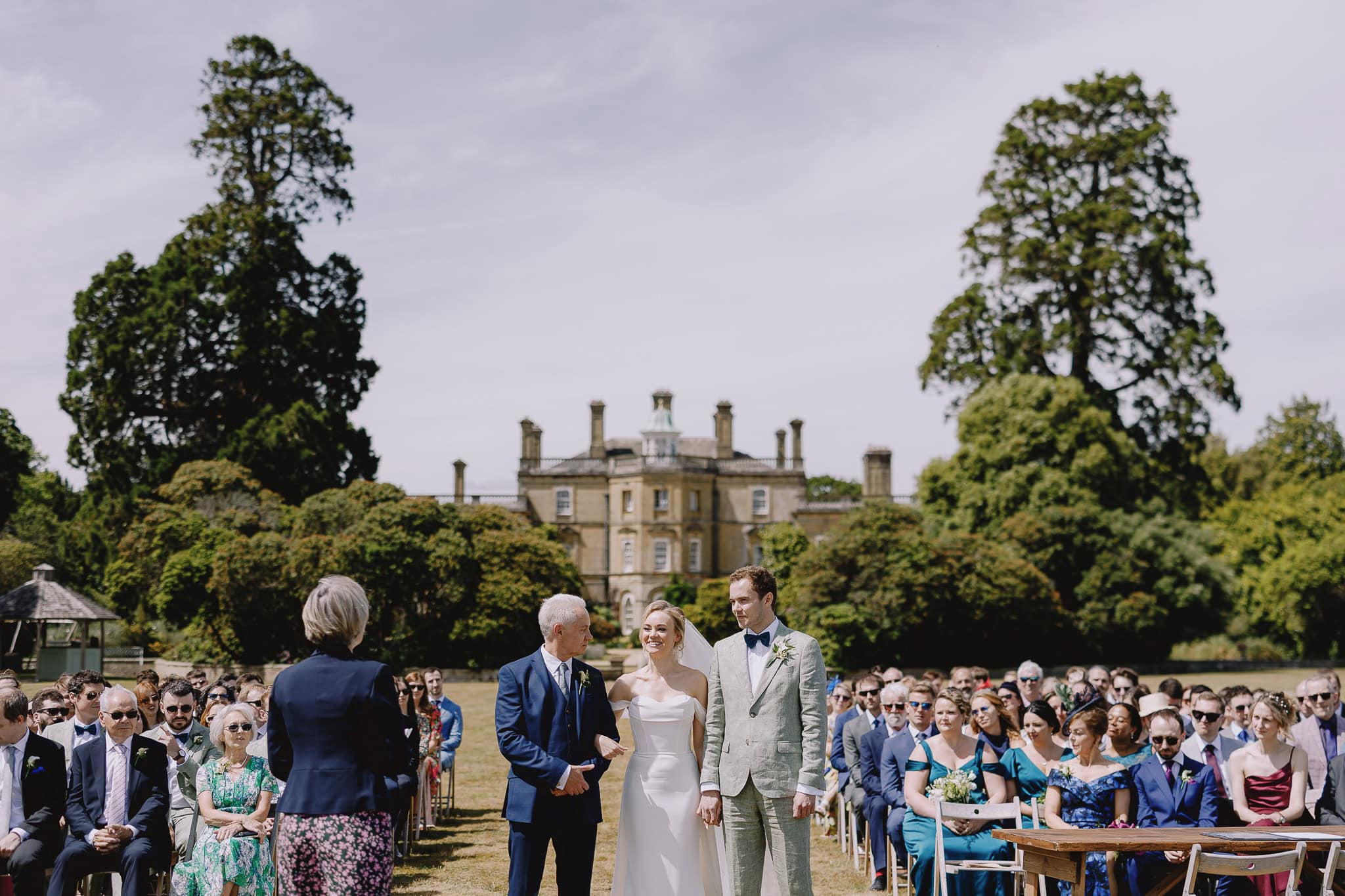 Outdoor wedding ceremony with bride, father, groom, and guests at a stately home.