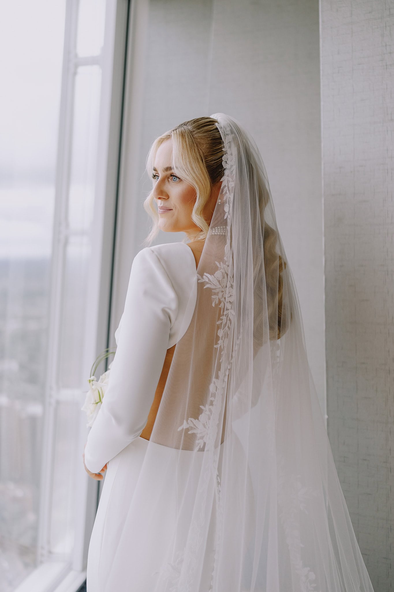 Bride in white gown and veil looking out window.