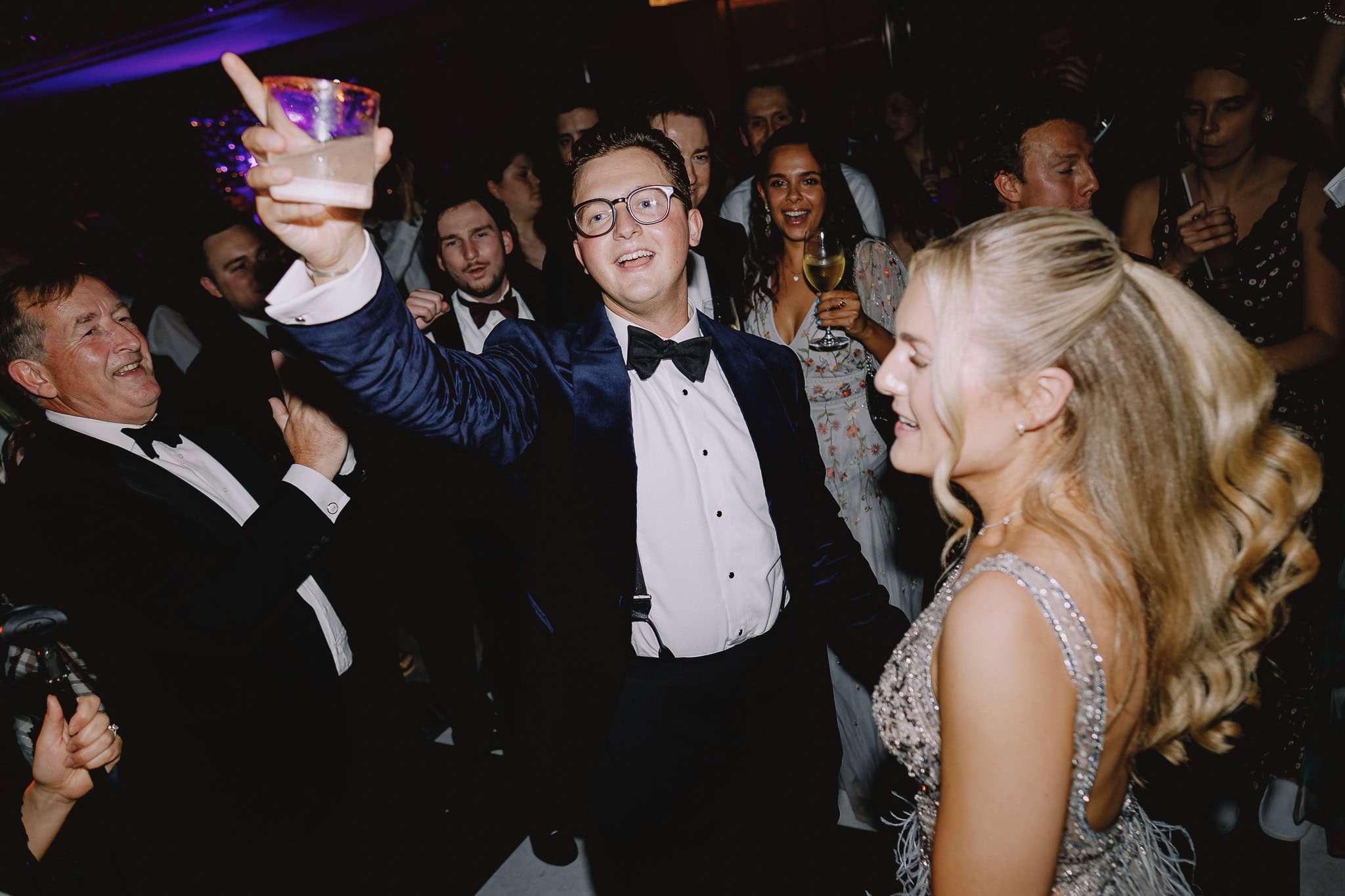 Festive party scene: man in tuxedo raises drink, surrounded by celebrating guests.