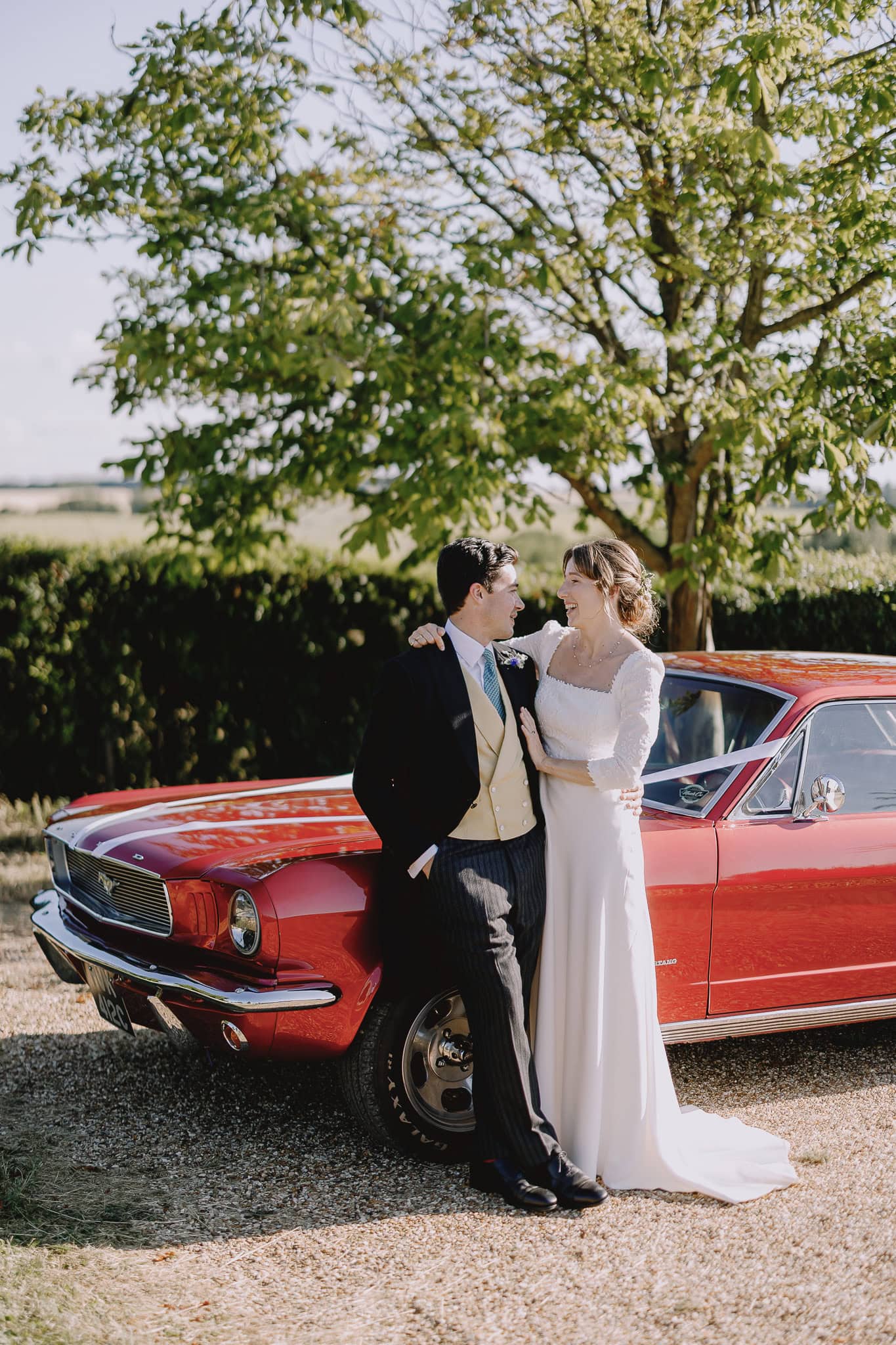 Wedding photo: Bride and groom pose with a red vintage Mustang car on their special day.