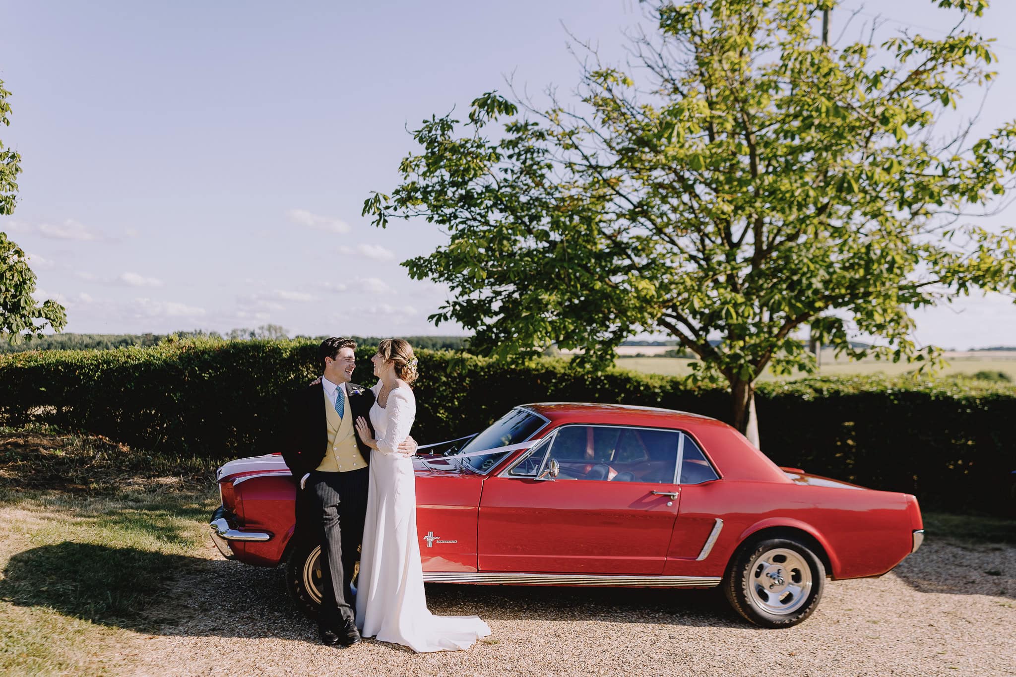Bride and groom embrace next to a vintage red Mustang on their wedding day.