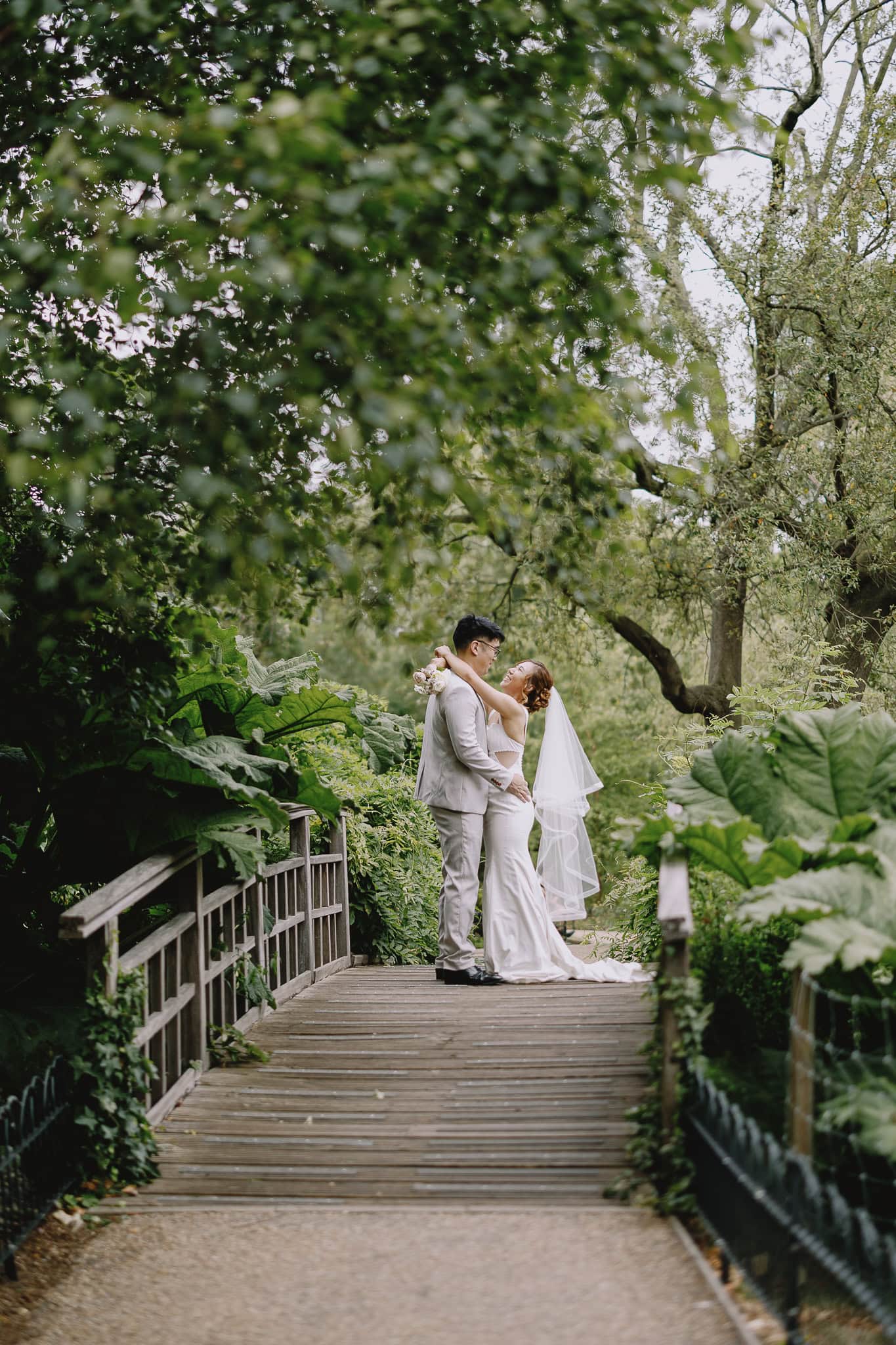 Bride and groom embrace on a wooden bridge in a lush green garden.
