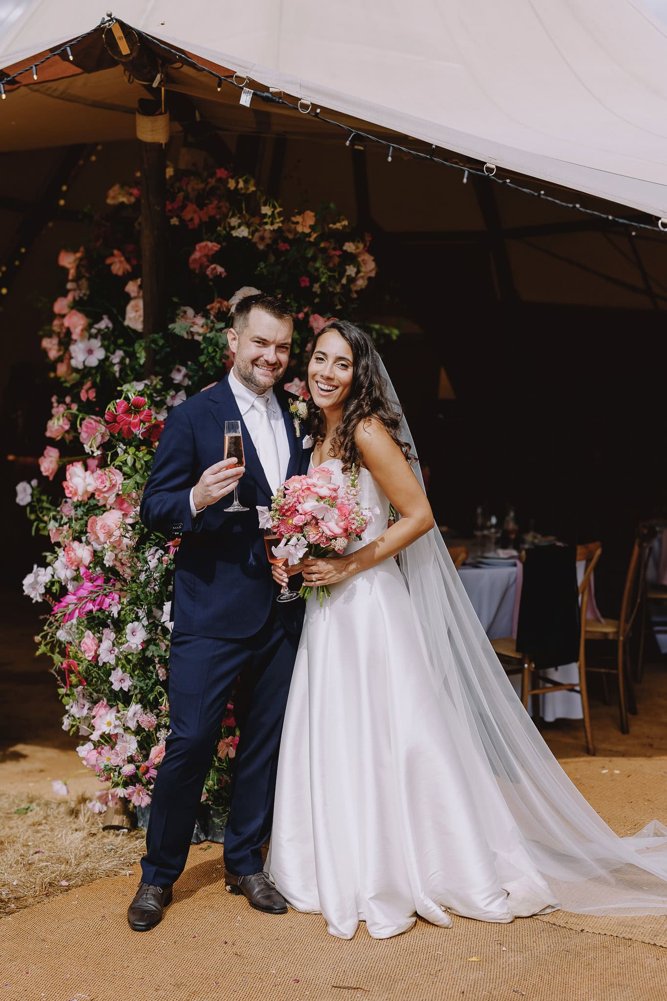 Happy bride and groom with champagne, posing by floral arch at outdoor wedding reception.