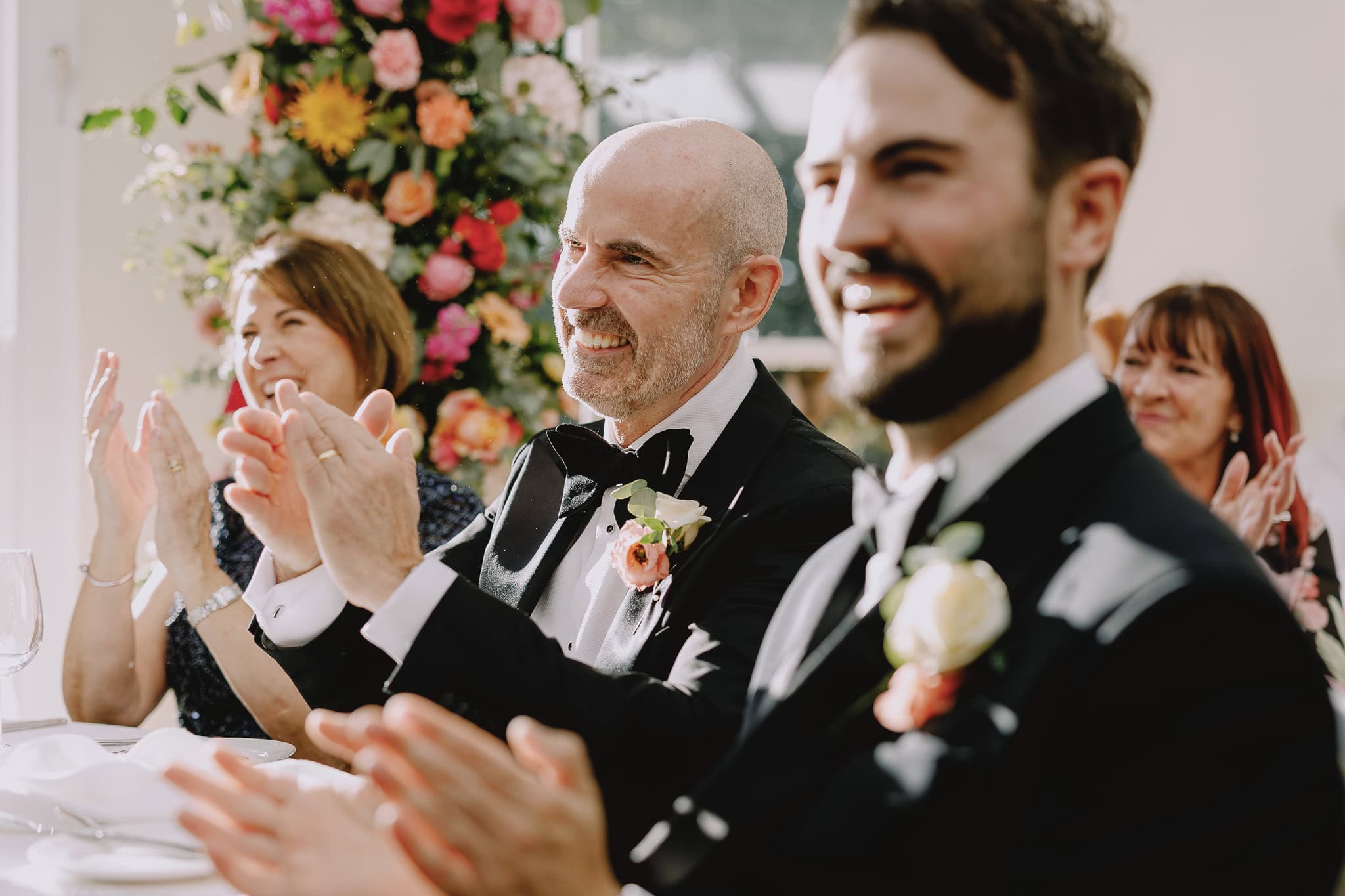 Wedding guests clapping, including men in tuxedos, at a reception.