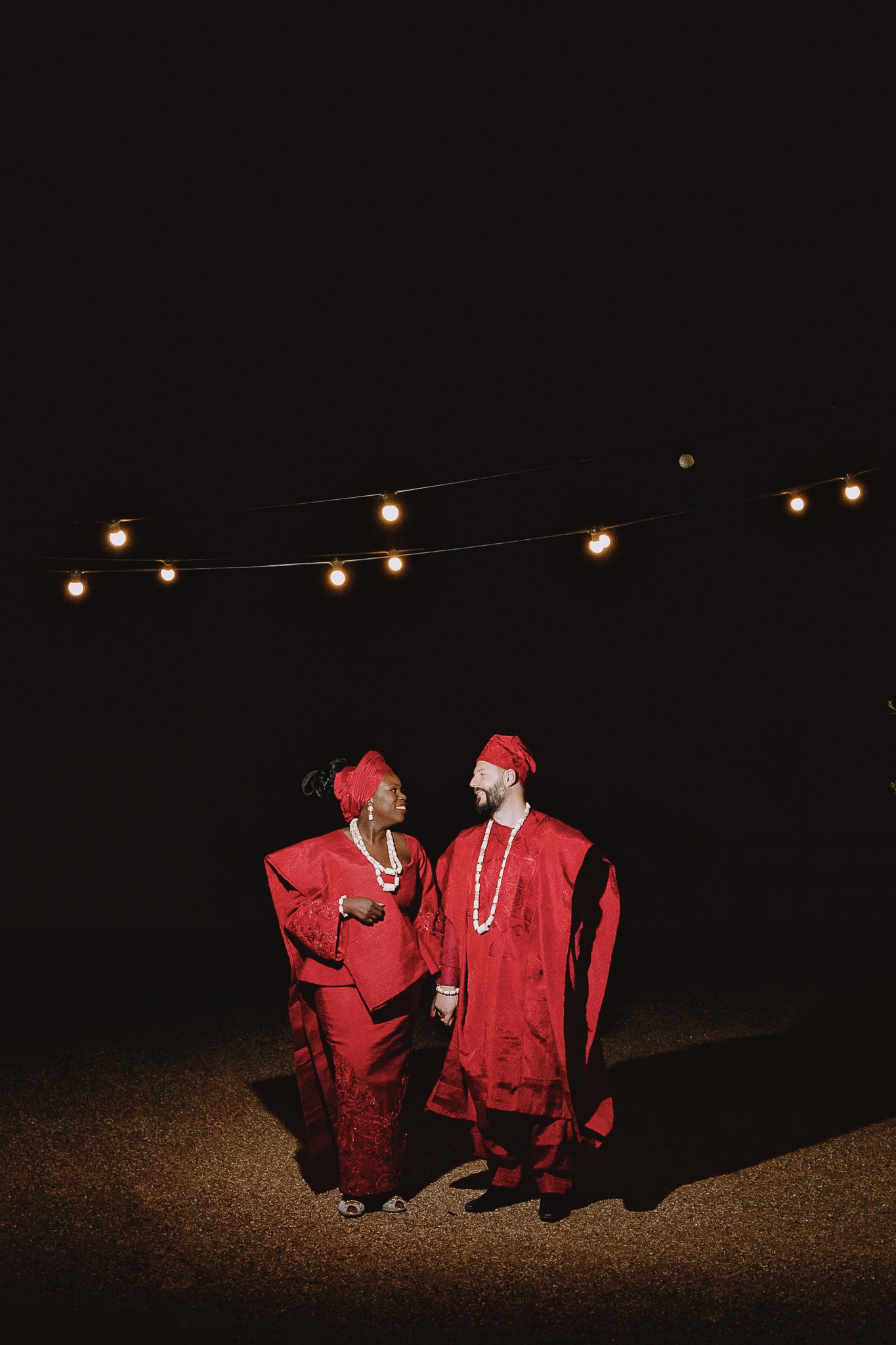 Couple in matching red traditional Nigerian attire holding hands under string lights.