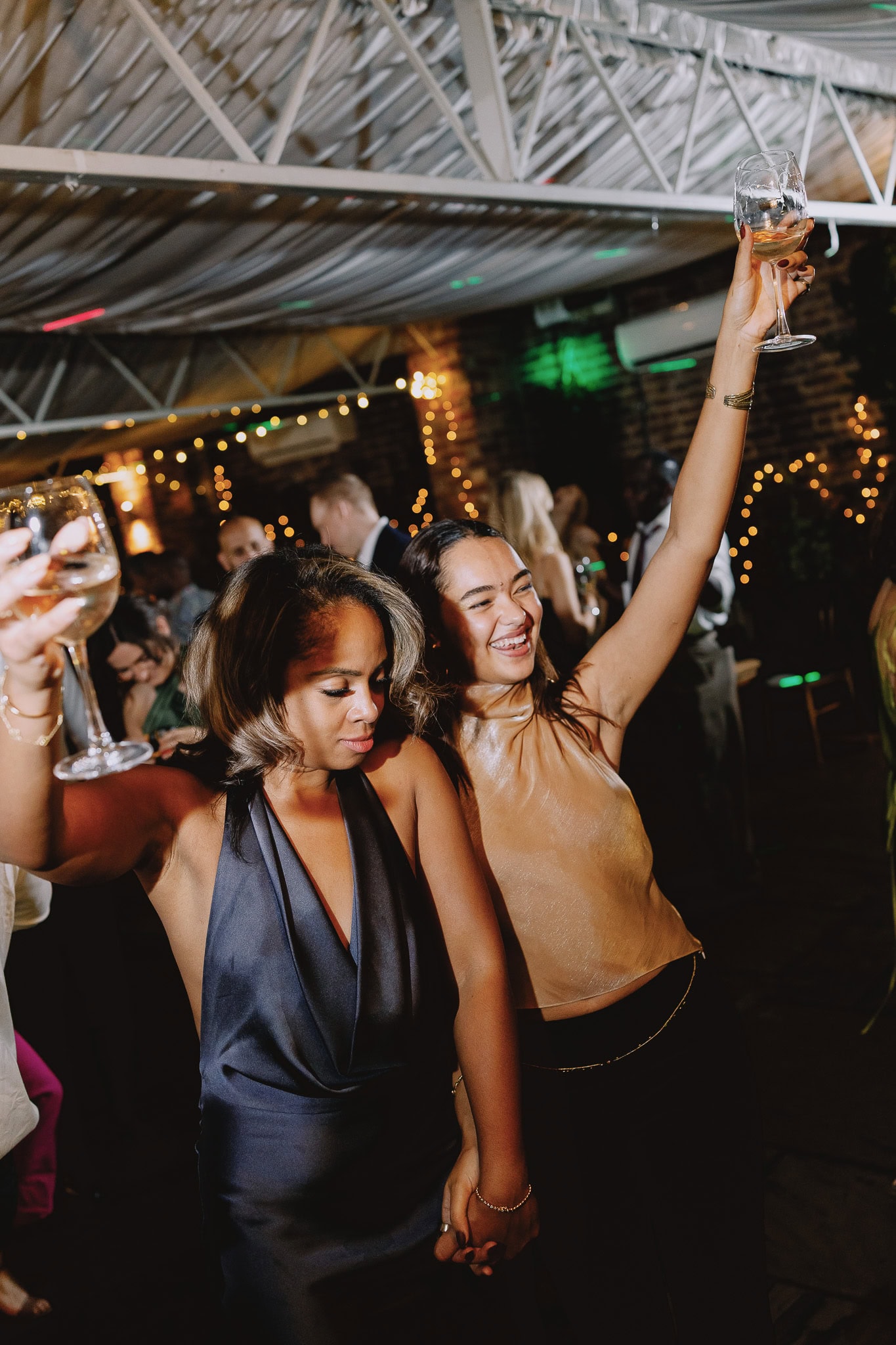 Two women dancing at a wedding reception, holding wine glasses and smiling.
