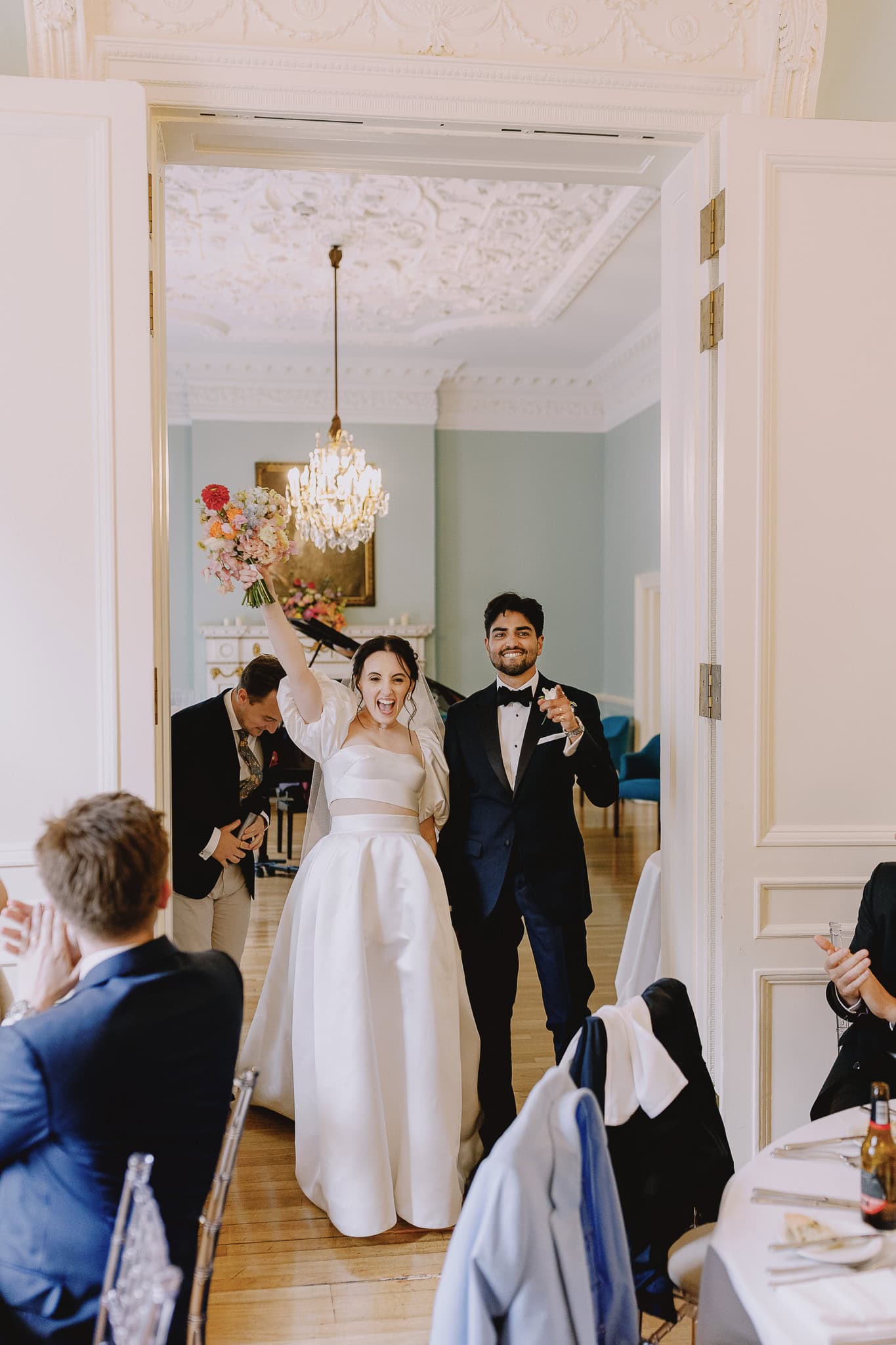 Newlyweds' joyful entrance at their wedding reception, chandelier overhead.