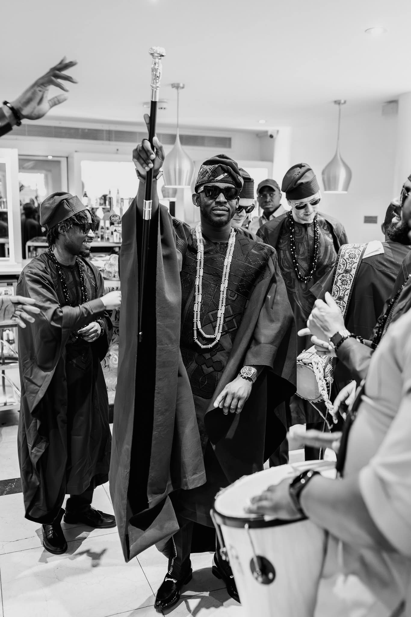 Man in traditional Nigerian Igbo attire holding a staff, surrounded by musicians.