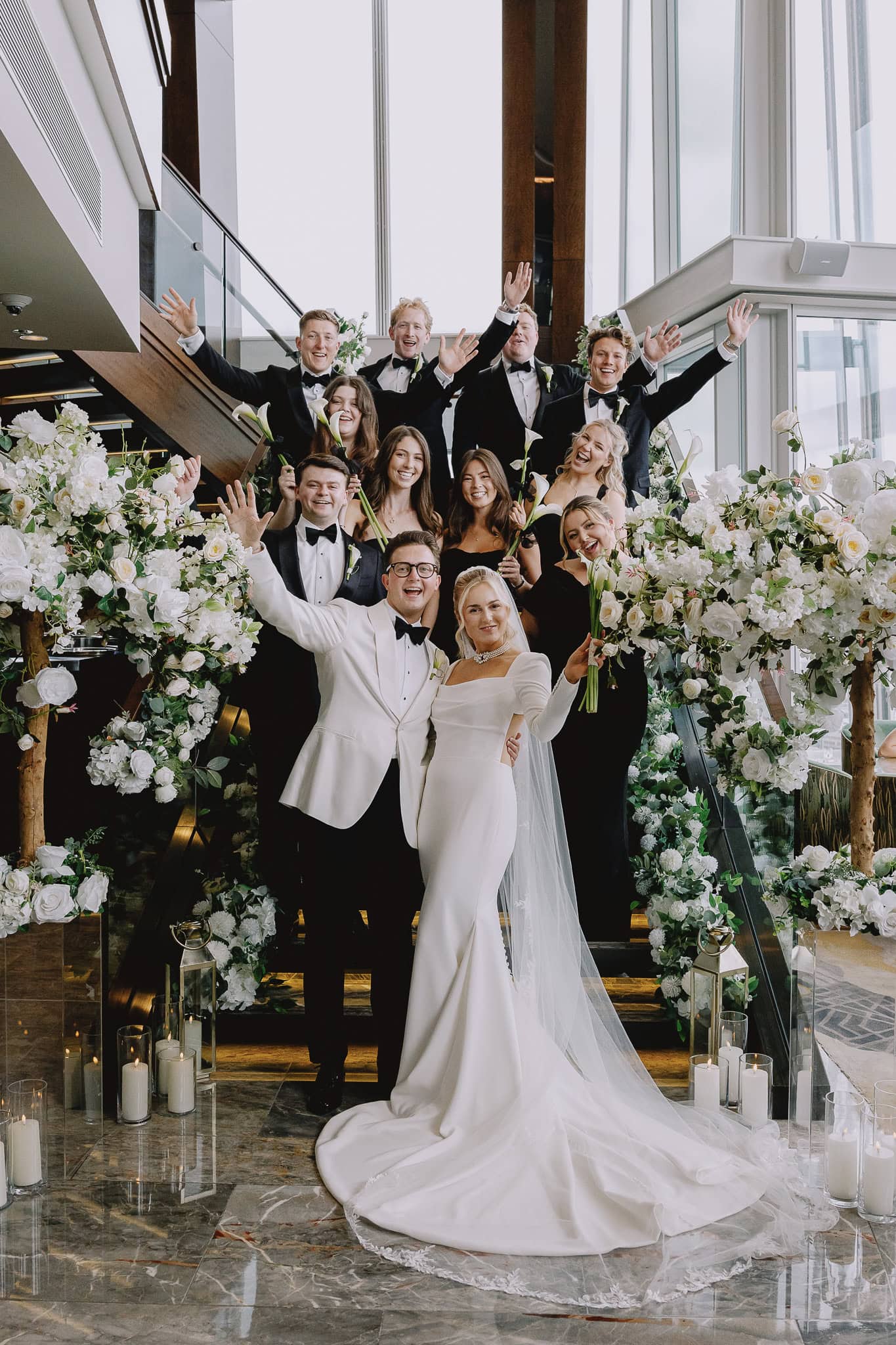 Wedding party on stairs with white floral decor, bride and groom in front.