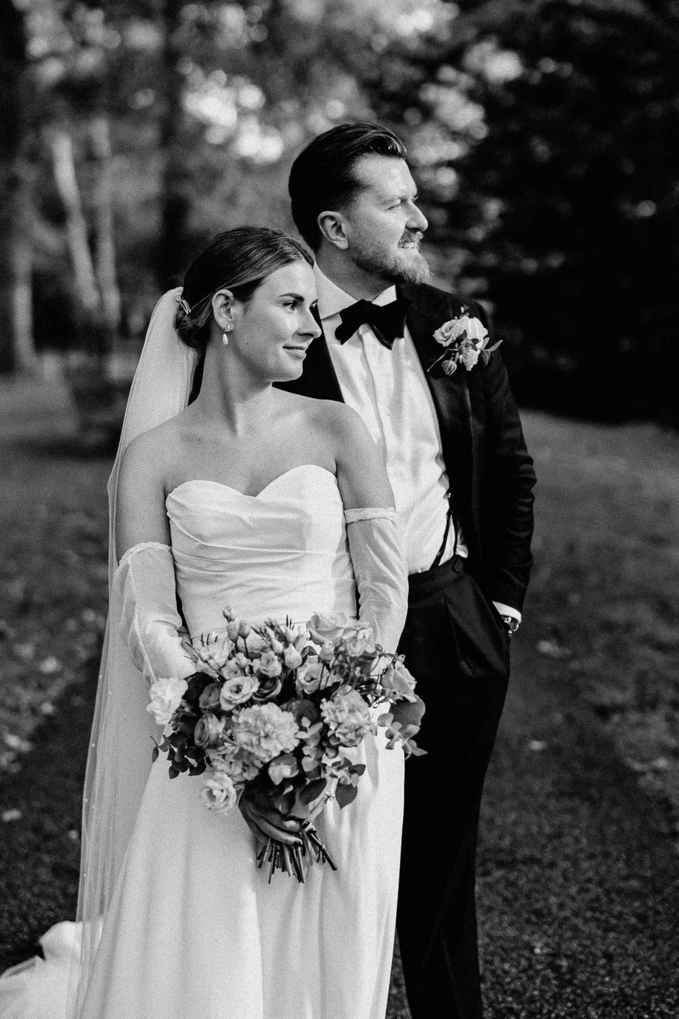 Bride and groom in wedding attire, looking off to the side with a bouquet.