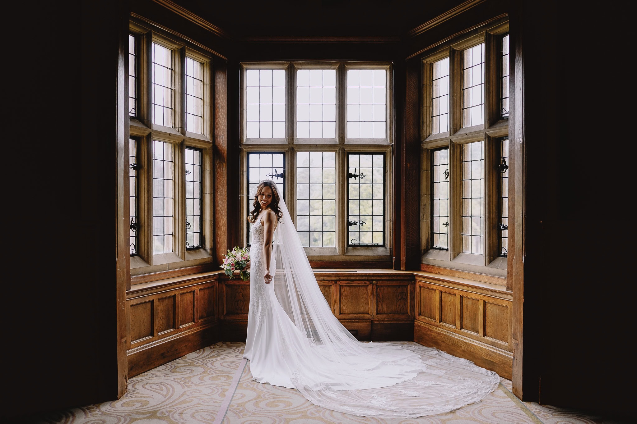 Bride in lace wedding dress stands in a wood-paneled room with large bay windows.