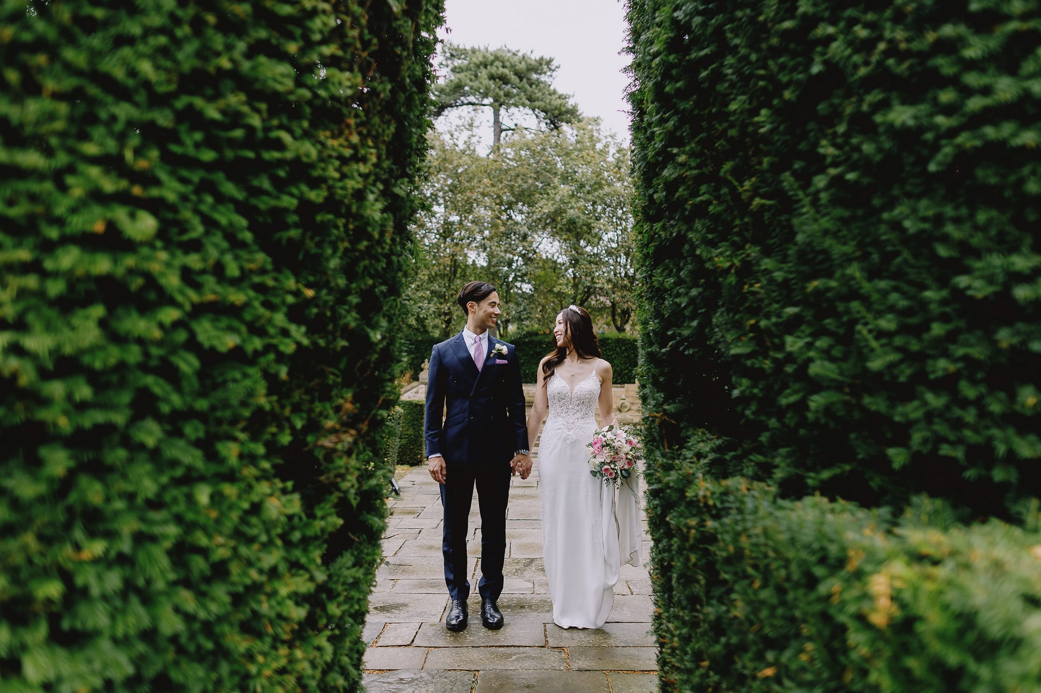 Bride and groom holding hands in a garden path framed by hedges. Wedding day portrait.