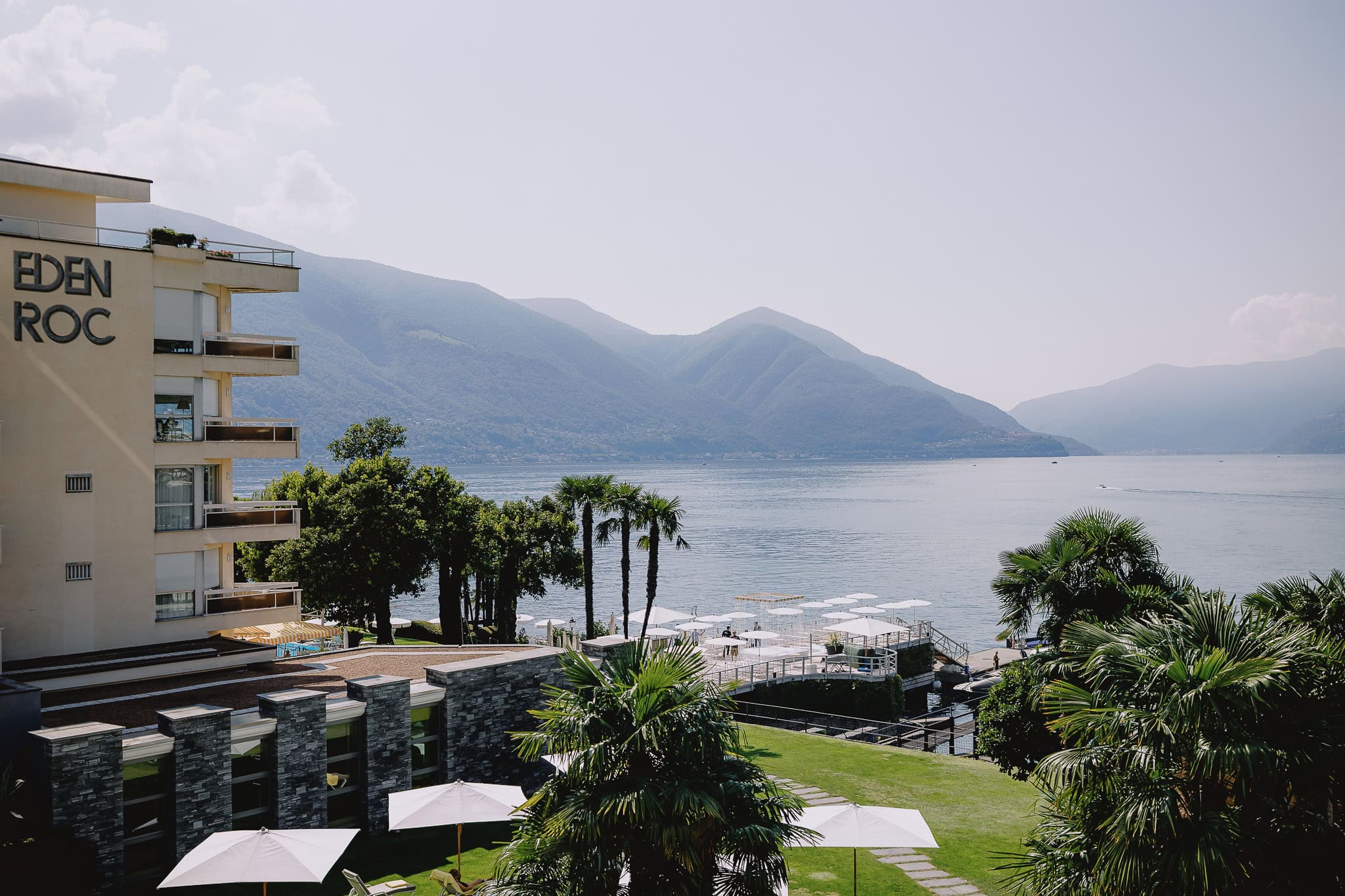 Eden Roc hotel overlooking Lake Maggiore, with mountains in the background. Palm trees and white umbrellas.