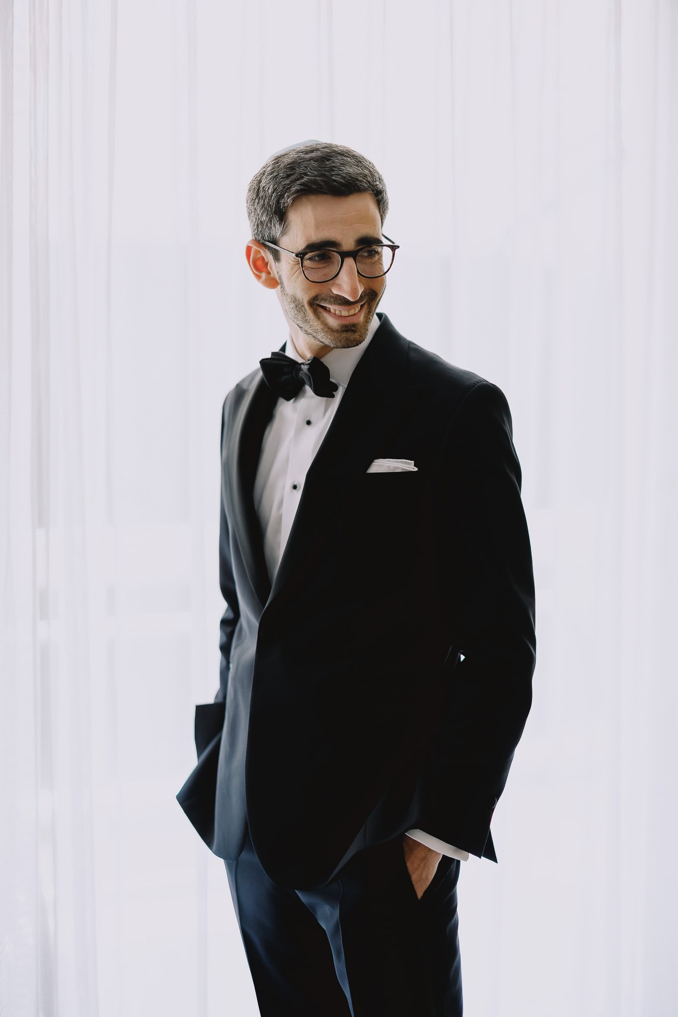 Groom in black tuxedo and bow tie smiles, looking to the side