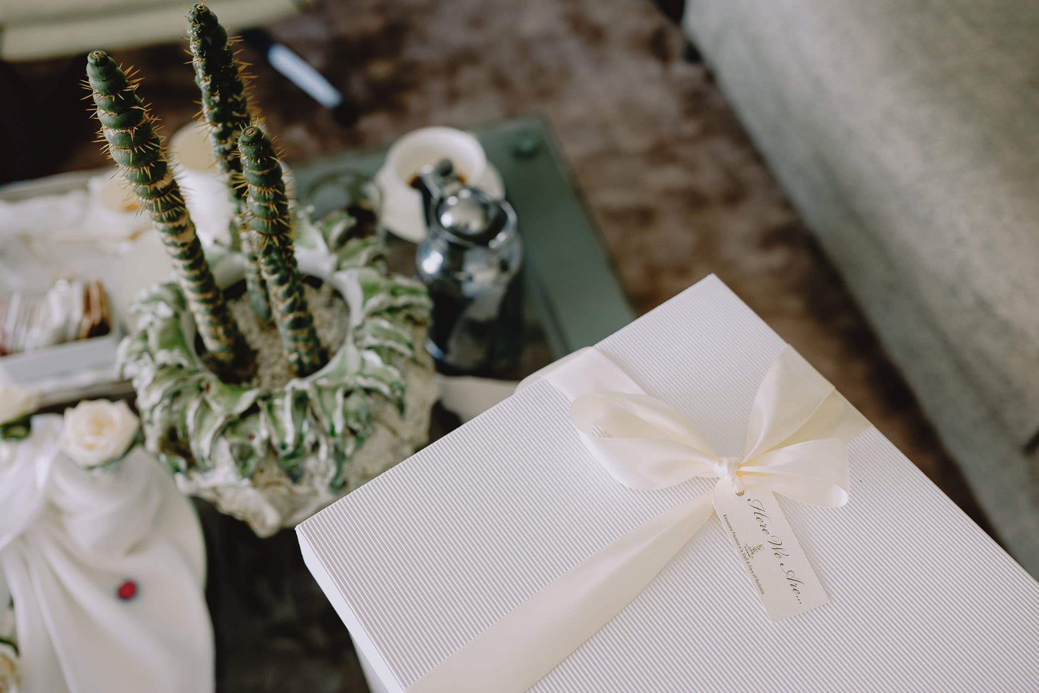 White gift box with ribbon, cactus plant, and tea set on a table. Text: "Here We Are".
