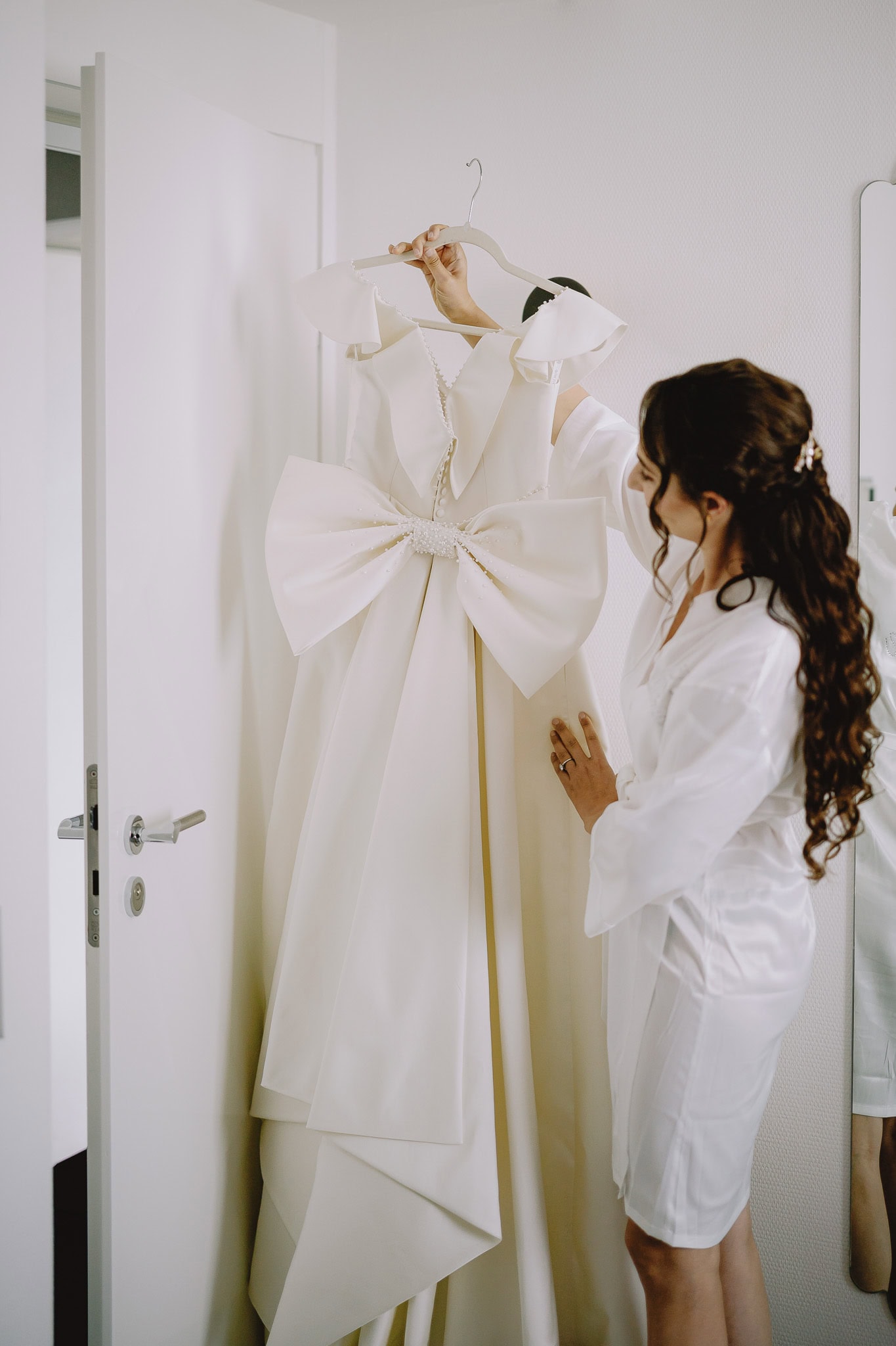 Bride in robe admires her elegant wedding dress with large bow detail.