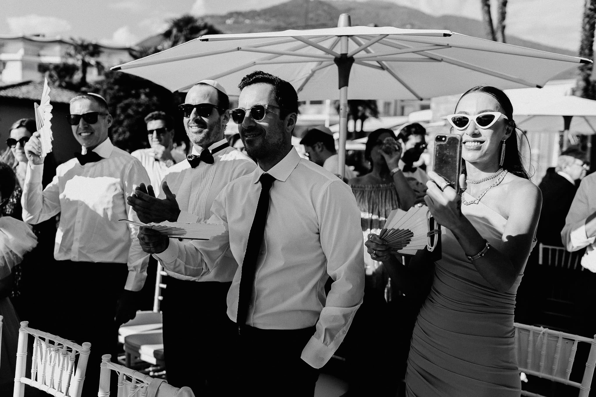 Wedding guests under umbrella, some holding fans, dressed in formal attire.