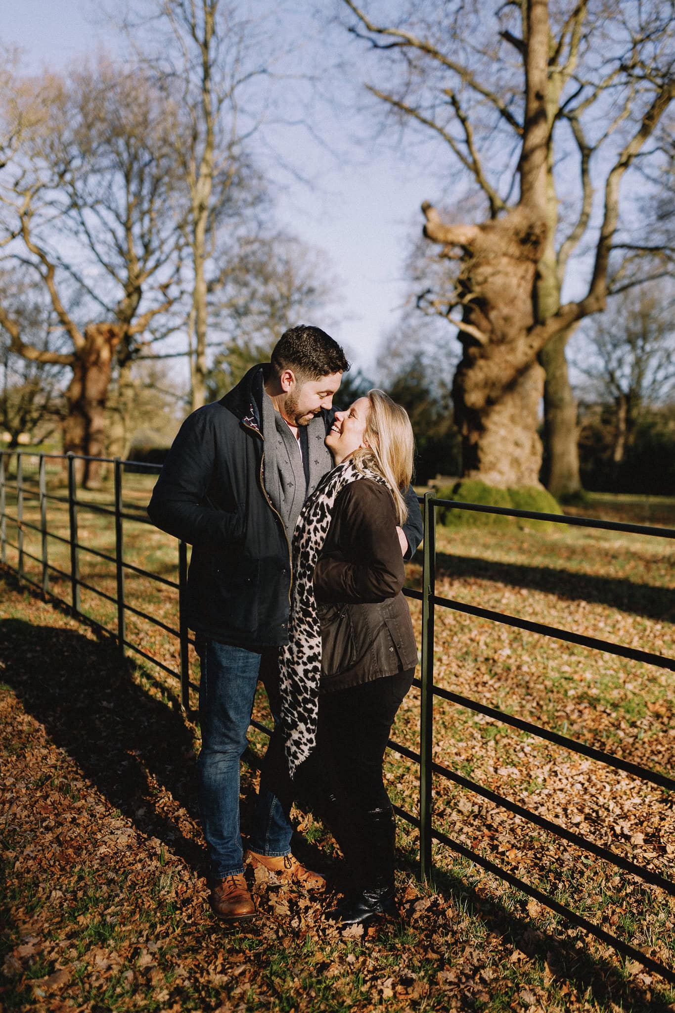 Loving couple embraces by a fence in a park, sunlight streaming through bare trees.