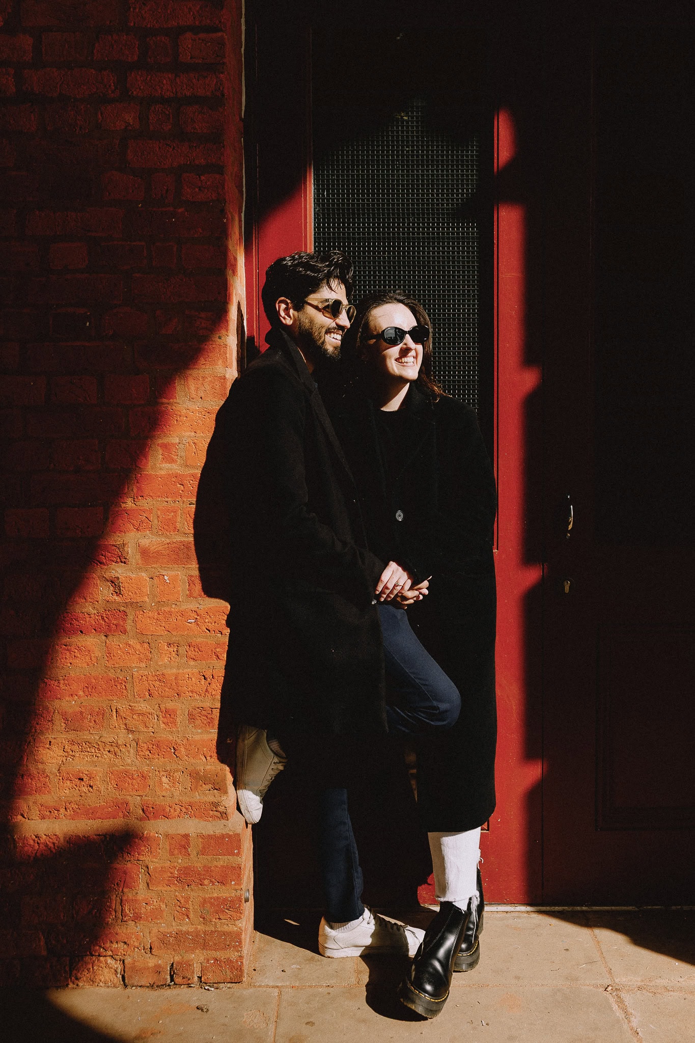 Couple in sunglasses leans against brick wall and red door, wearing black coats and stylish shoes.