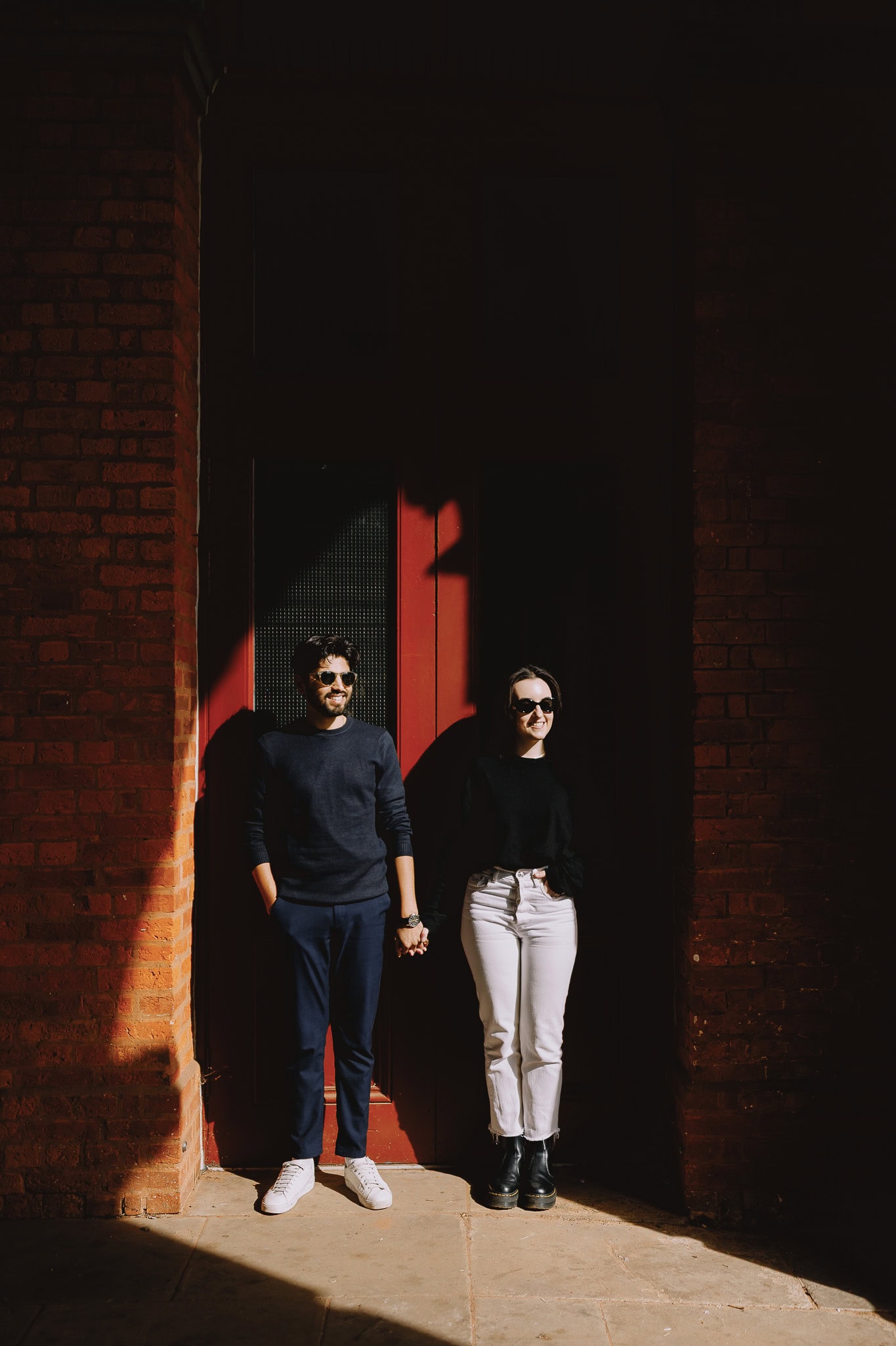 Couple holding hands in front of a red brick building, smiling in the sunlight.