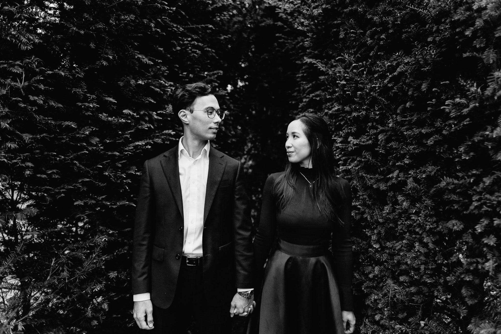 Couple holding hands, looking at each other in front of greenery. Romantic engagement photo.