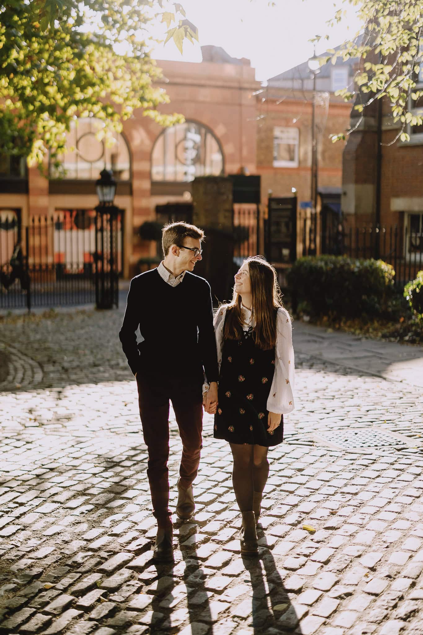 Couple walking hand-in-hand on a cobblestone street, smiling at each other in golden sunlight.
