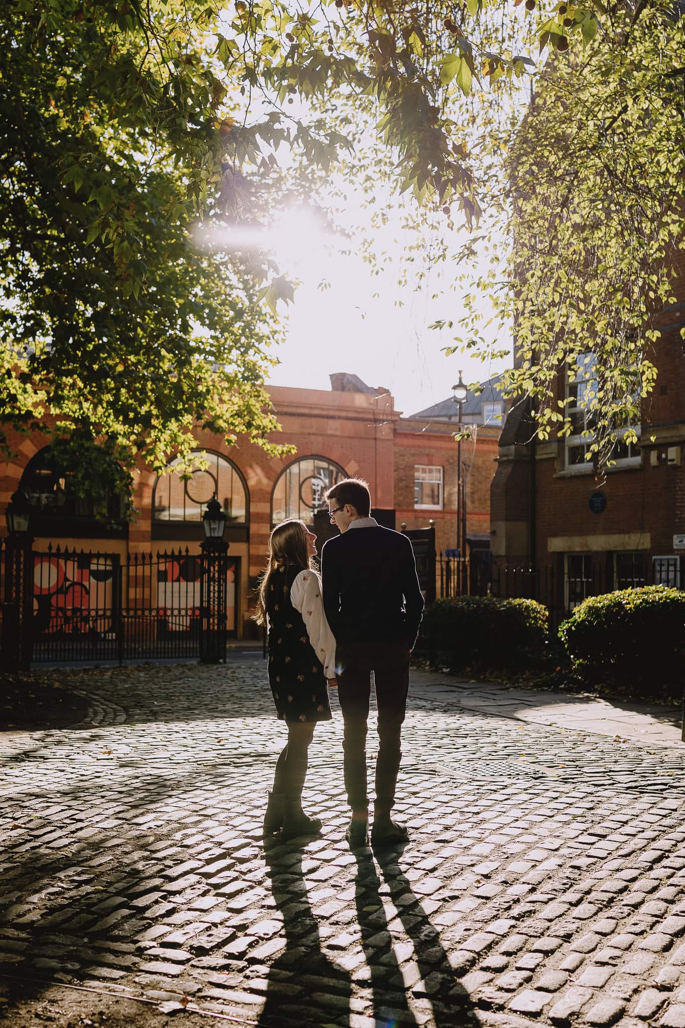 Couple silhouetted in golden sunlight, walking on cobblestone street.