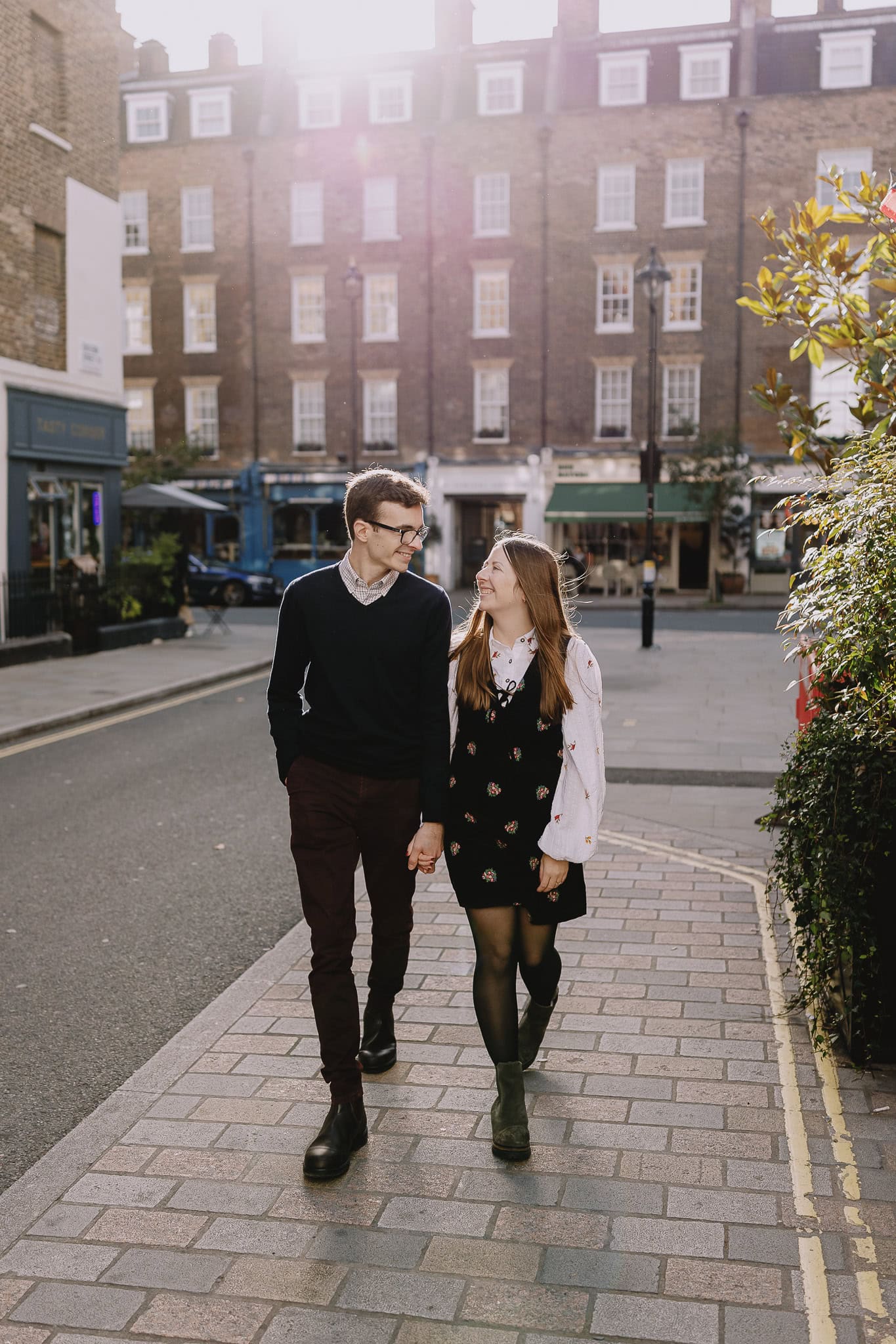 Couple holding hands, walking on a cobblestone street in London. Smiling at each other.