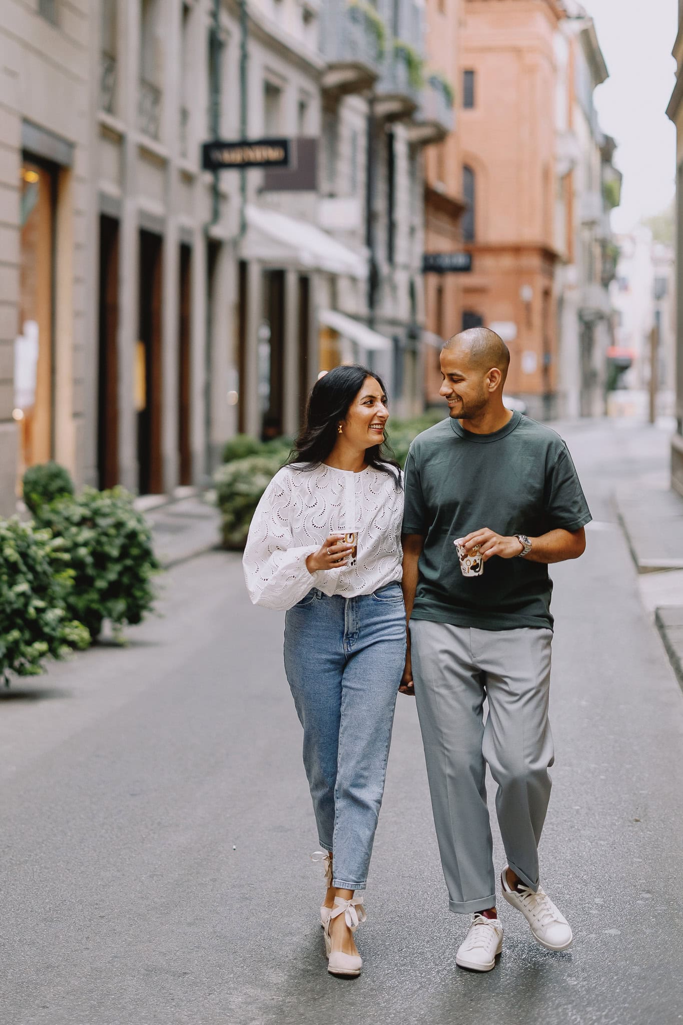 Couple walking on a European street, holding drinks and smiling at each other.