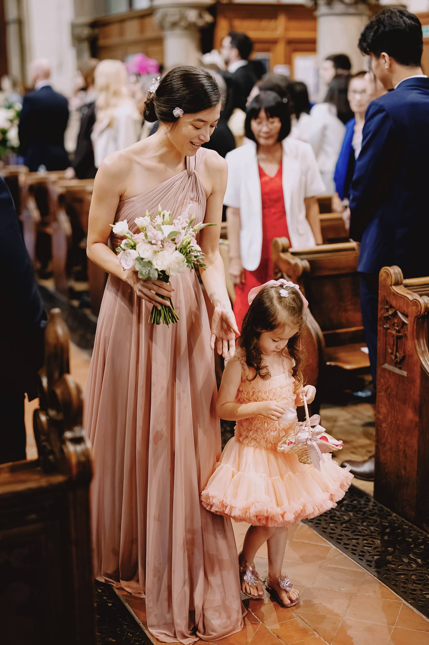 Bridesmaid guides flower girl down aisle, both in peach dresses.