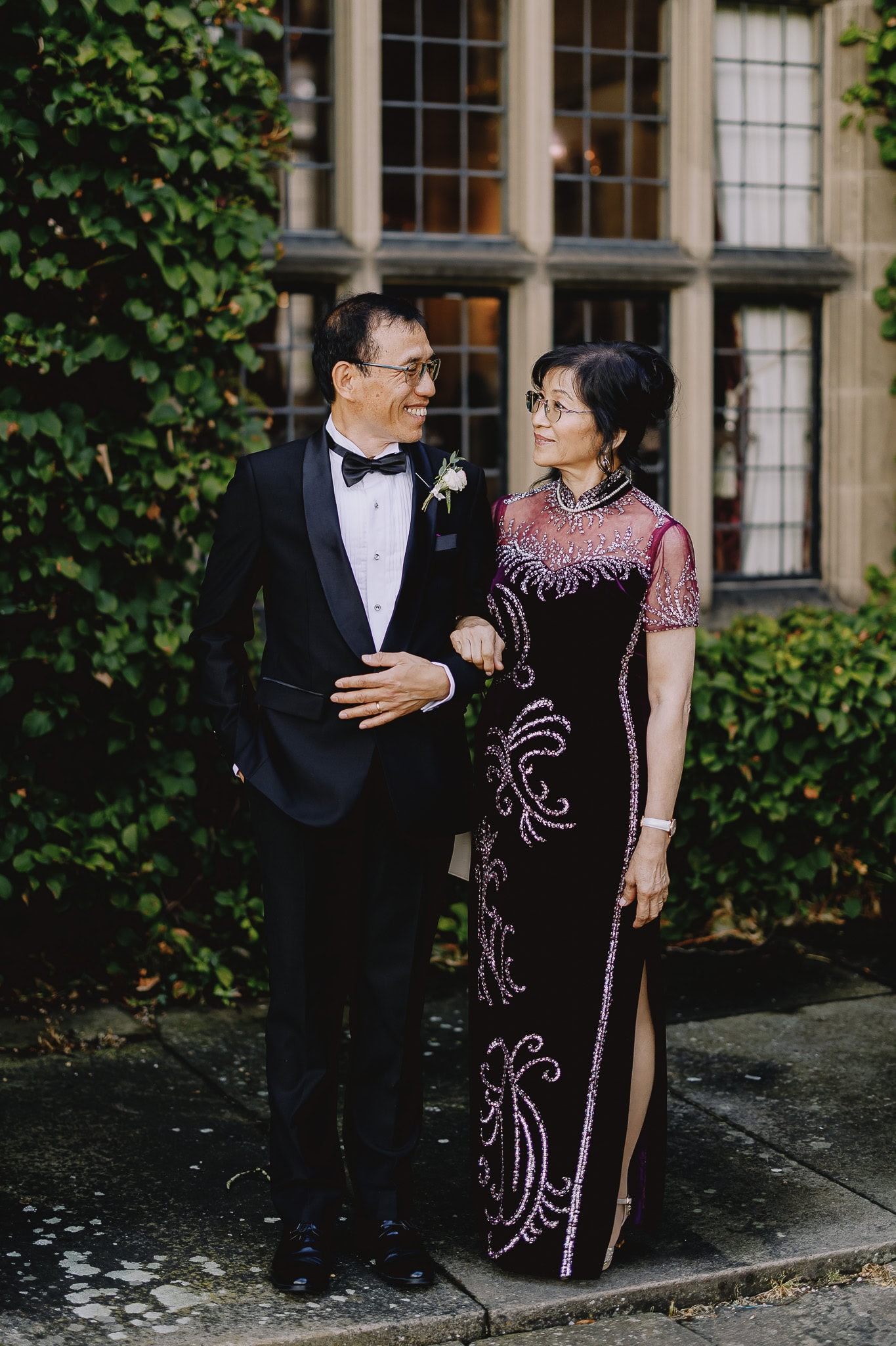 Elegant Asian couple in formal wear posing outdoors by ivy-covered building.