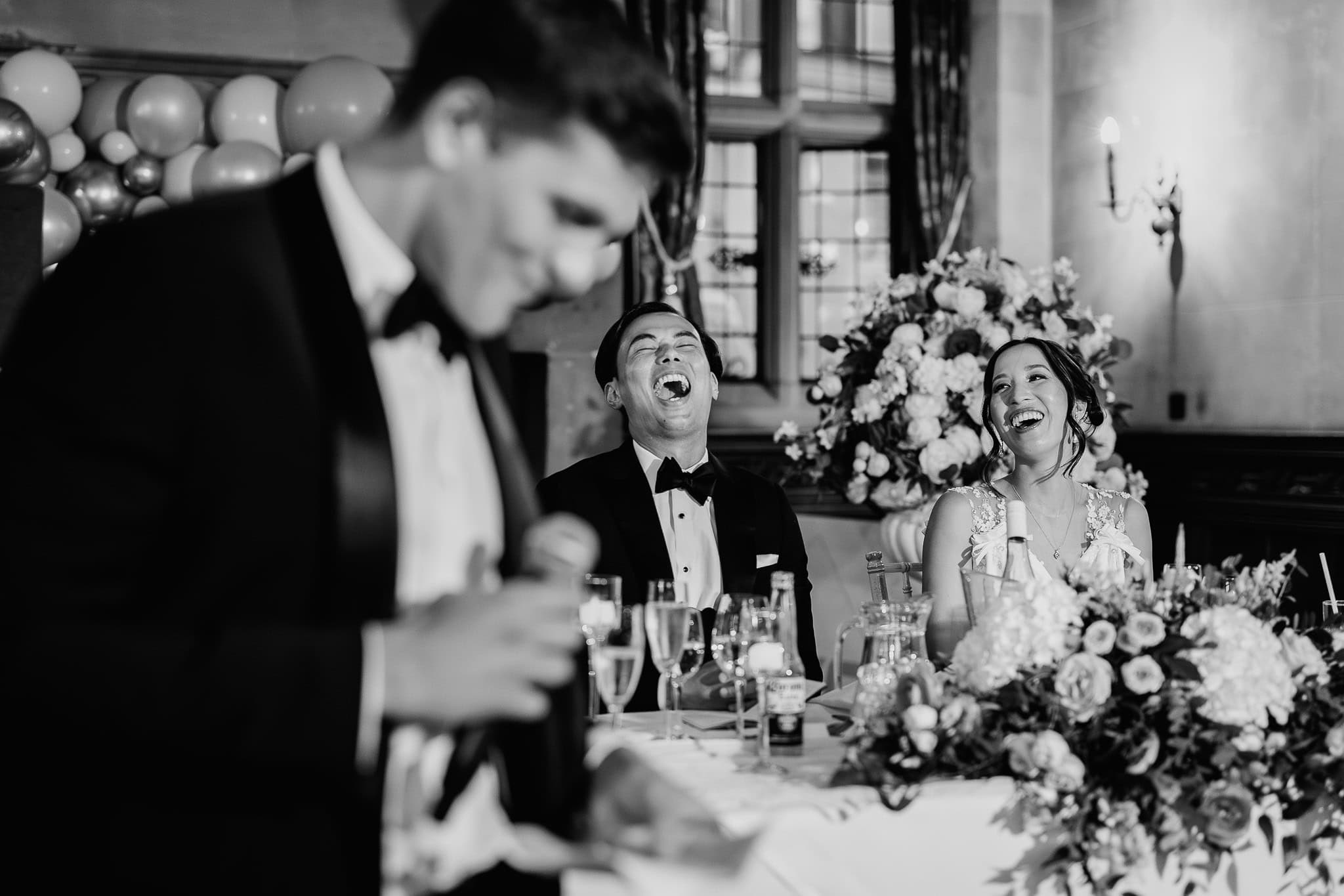 Groom laughing during a wedding speech, bride smiling beside him at the reception.