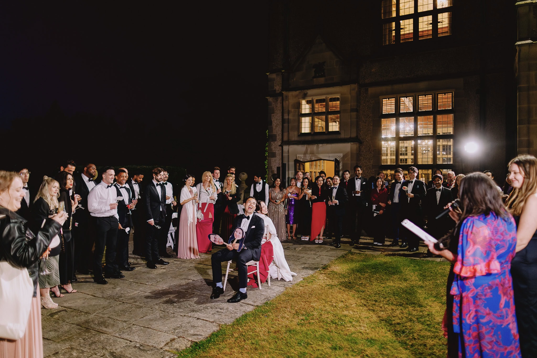 Wedding guests cheer as groom holds a cutout of bride, standing in front of a large building at night