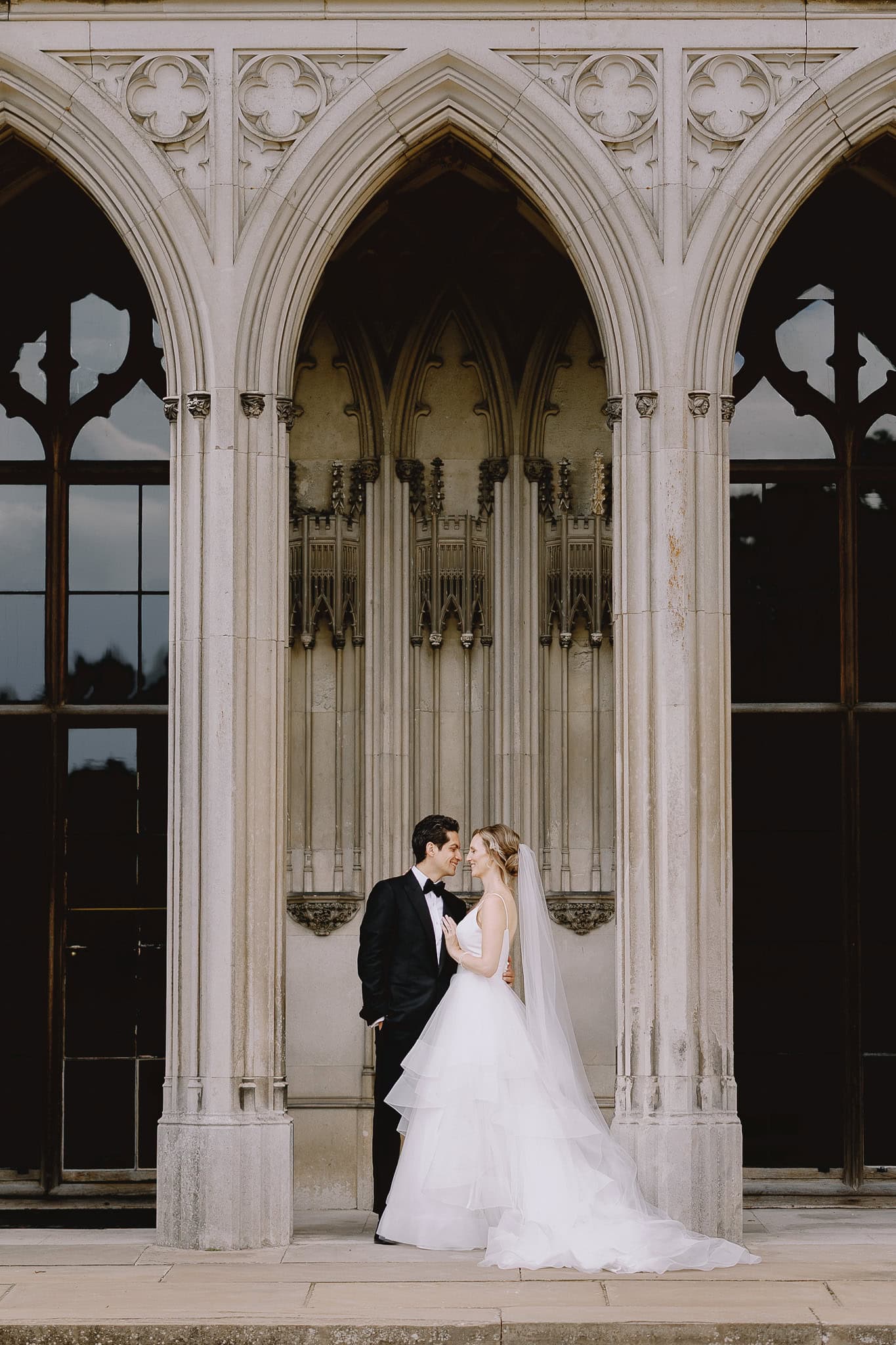 Editorial wedding photography: Bride and groom embrace in front of gothic architecture.